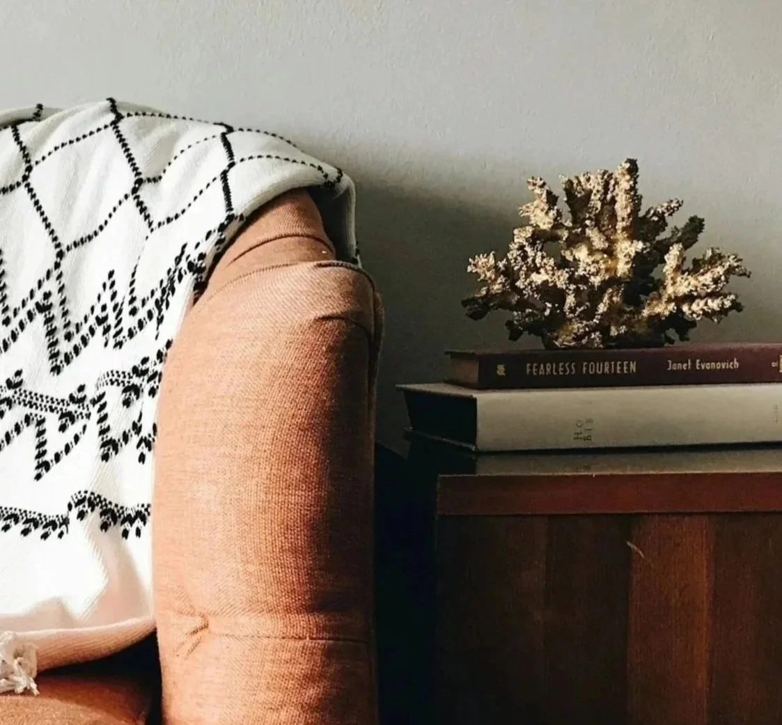 Close-up of a wooden side table with a large book titled "Fearless Fourteen" by Janet Evanovich, a decorative coral piece, and a stack of other books, next to a mustard yellow upholstered armchair with a black and white patterned throw blanket draped over its back.
