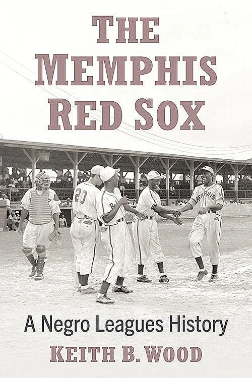 Black and white photo of young African American baseball players on a field, with some shaking hands and others standing nearby, under a covered bleachers.