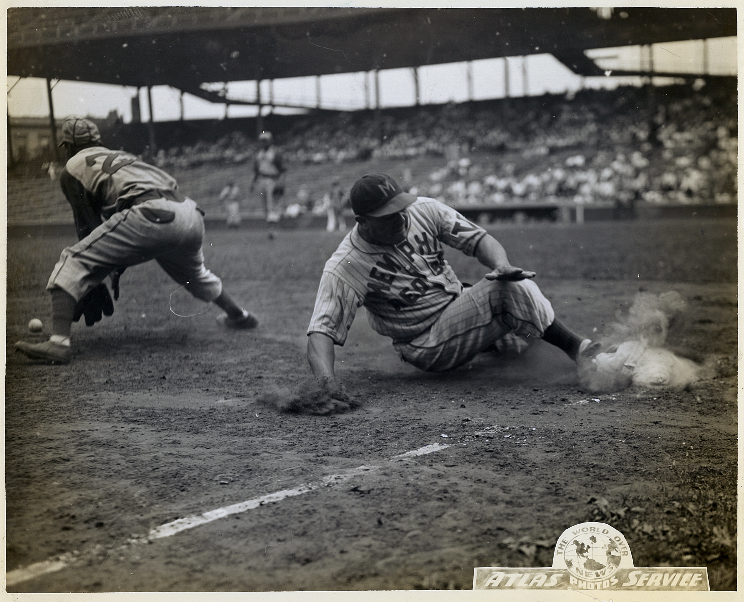 A black and white action photograph of a baseball game showing a player sliding into a base while a fielder is trying to catch the ball, with a crowd in the background.