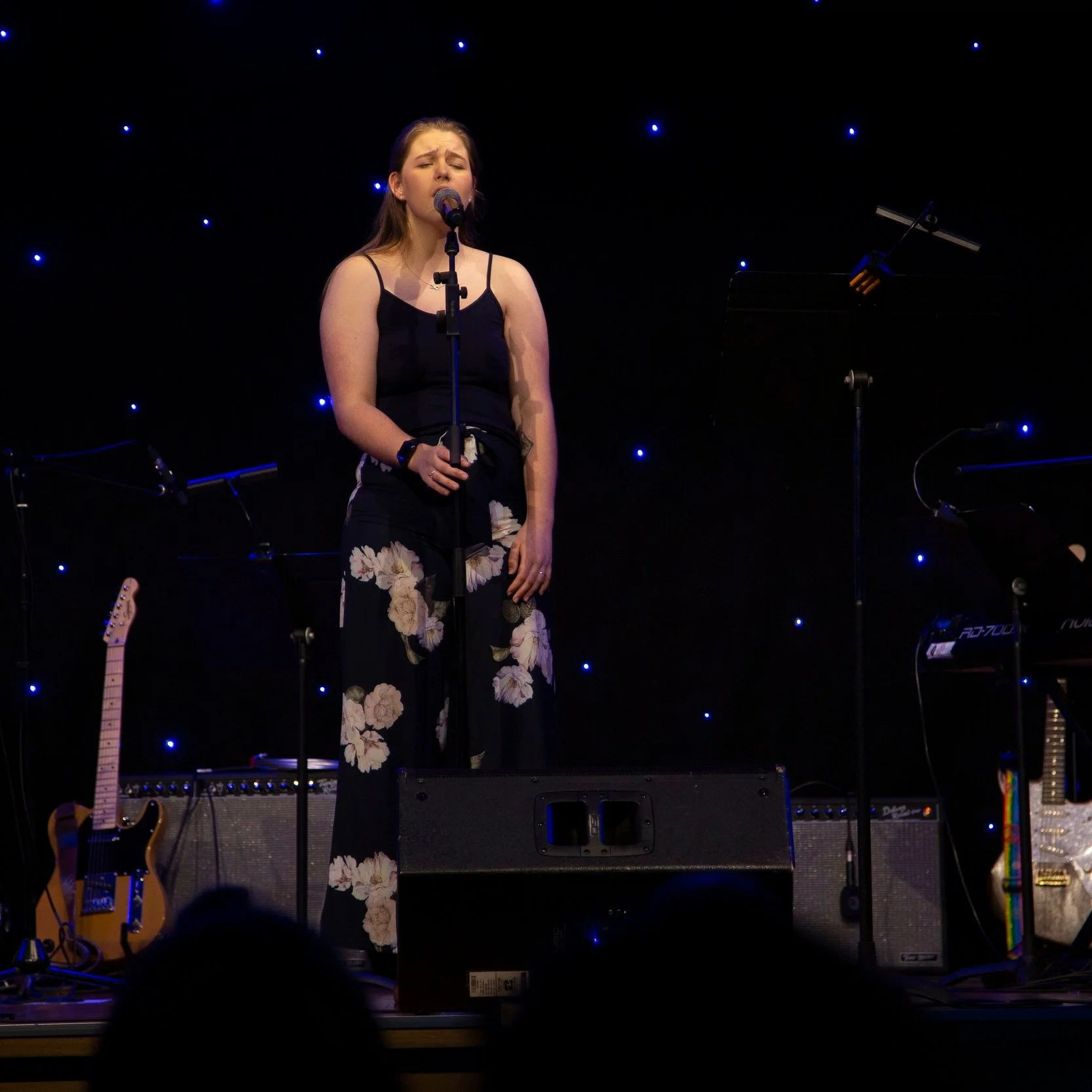 Ella singing on stage with blue star-like lights in the background, accompanied by musical instruments including guitars and a keyboard.