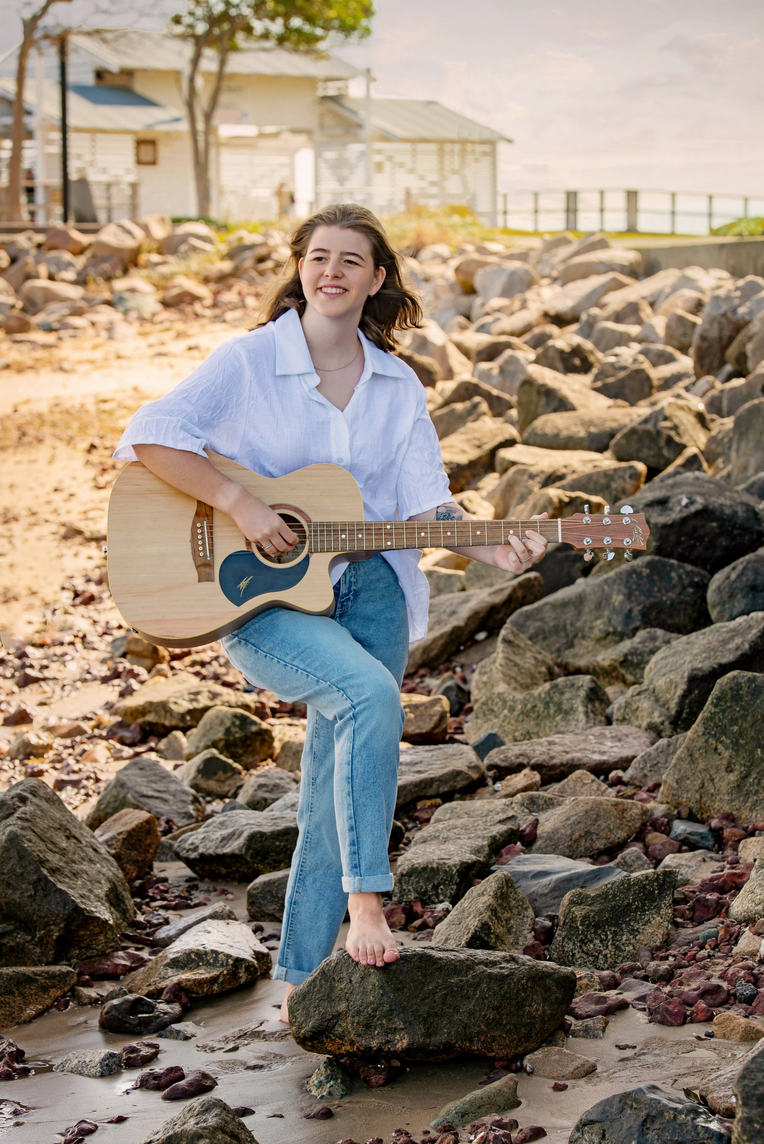 A young woman named Ella with long brown hair playing an acoustic guitar while barefoot on rocks by the beach, smiling and looking away.