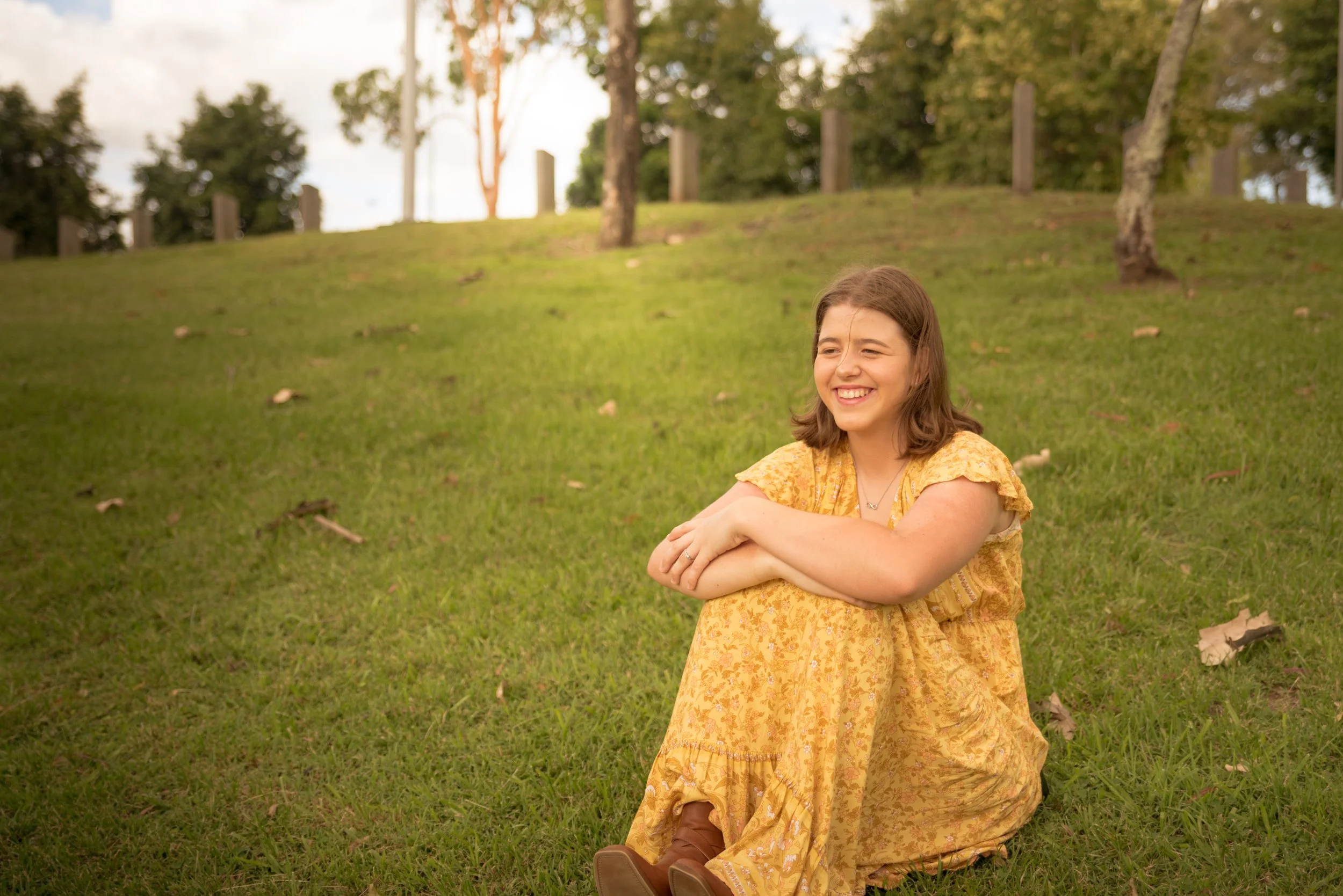 Ella sitting on the grass in a park, smiling and wearing a yellow dress, with trees and a grassy hill in the background.