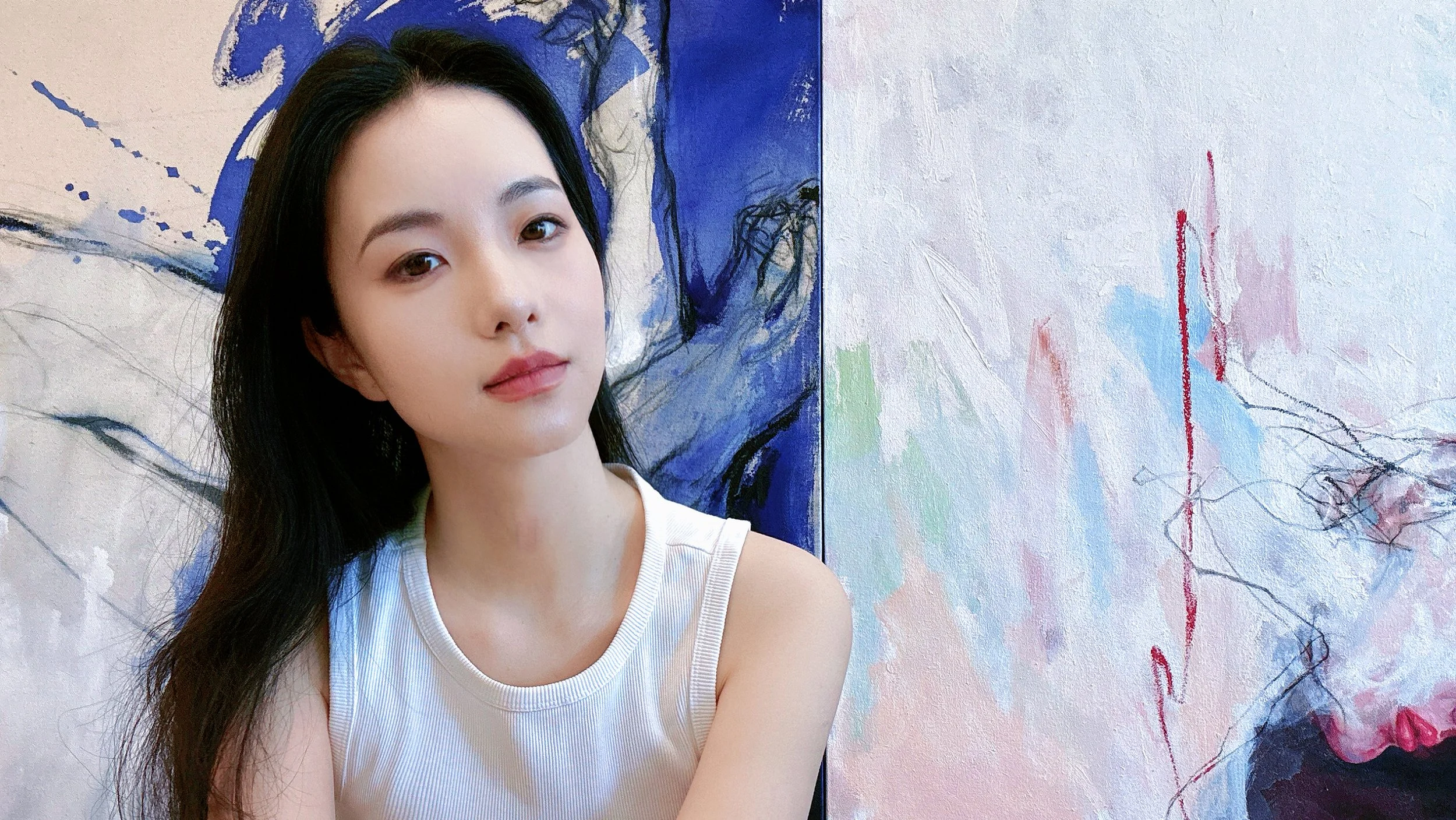 A woman with long black hair and fair skin posing in front of a colorful abstract painting with blue, pink, and black strokes.