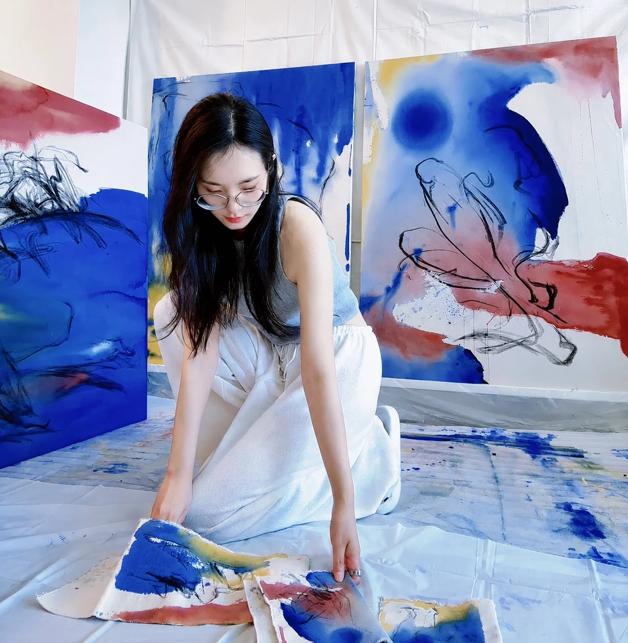 A woman with long dark hair and glasses kneeling on the floor in front of abstract watercolor paintings in blue, red, yellow, and white. She is holding a piece of fabric with similar colors.