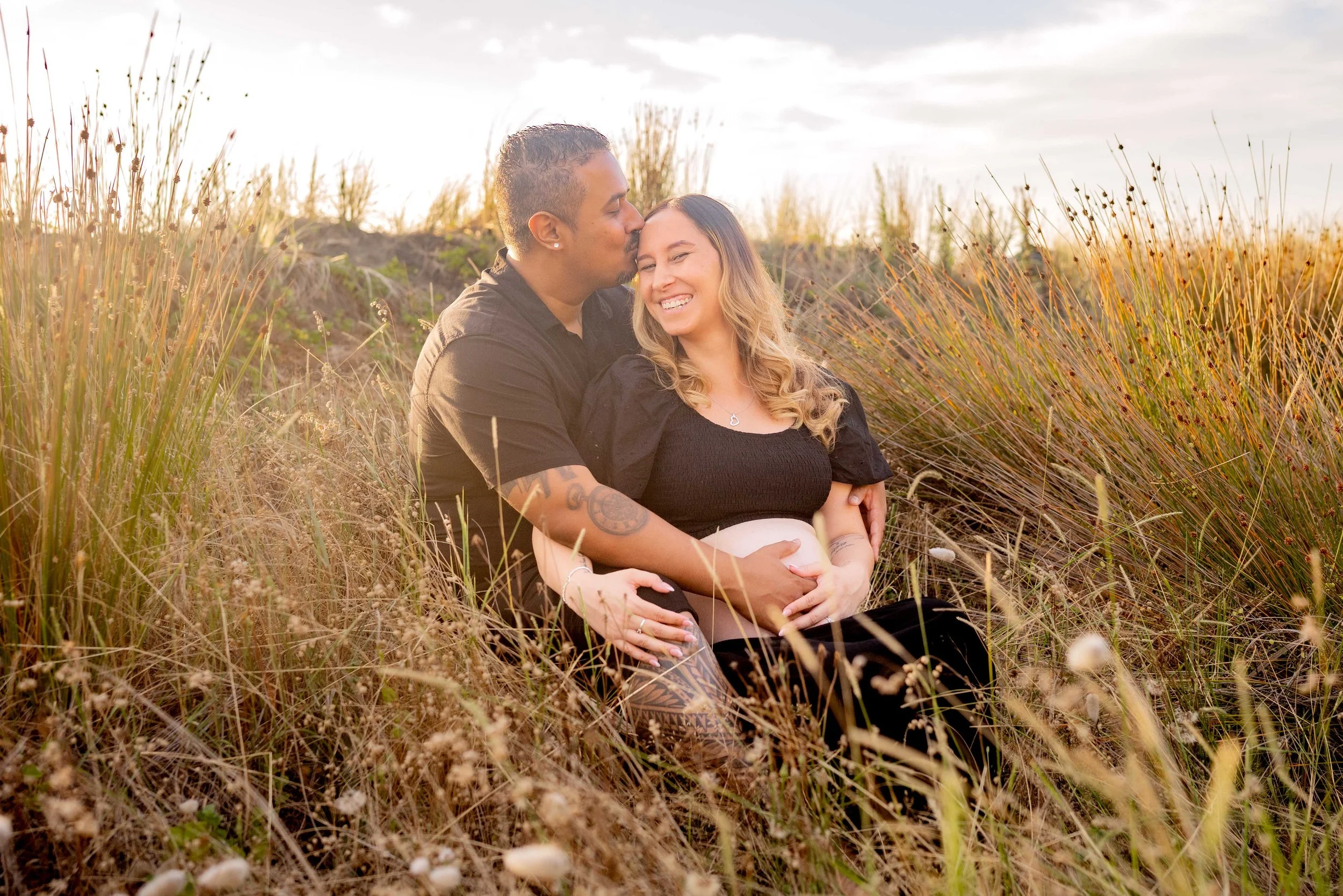 couples maternity photoshoot, in the sand dunes in auckland, he is kissing her forehead, she has a big smile on her face