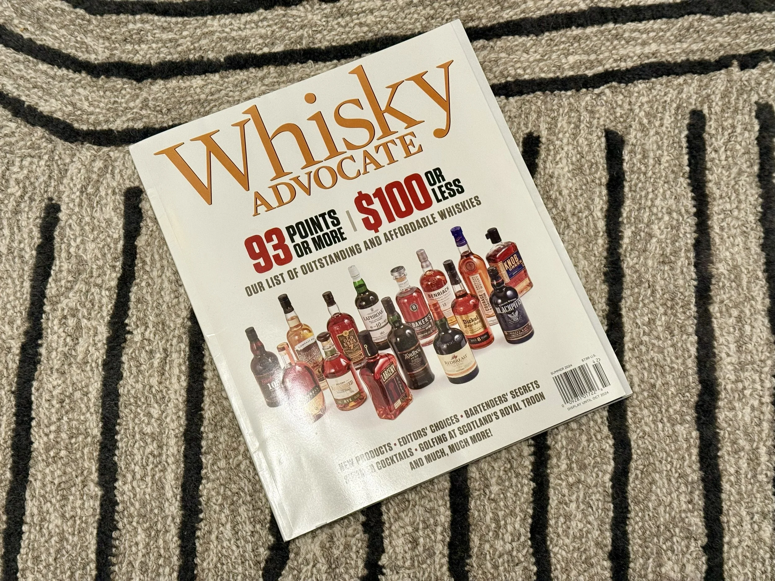 A magazine titled 'Whisky Advocate' on a striped textured carpet. The cover features images of various whiskey bottles and text promoting a list of outstanding whiskies priced at $100 or less, with a points system of 93 points or more.