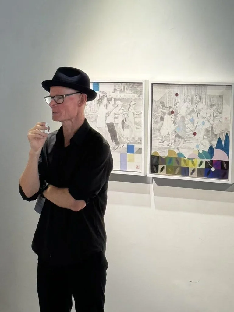 A man wearing a black hat and black shirt stands in an art gallery speaking into a microphone.