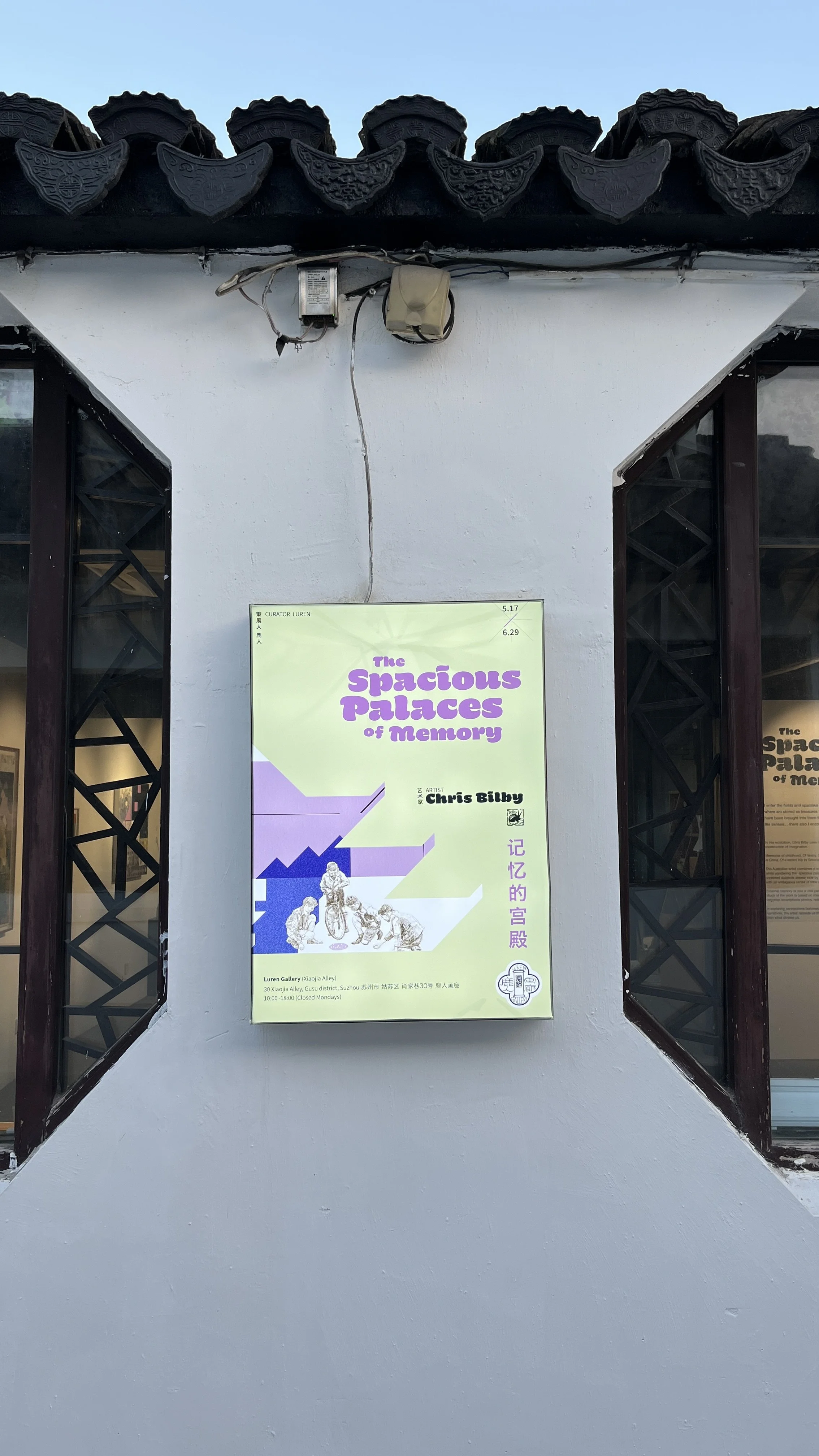 Poster for an art exhibition titled "The Spacious Palaces of Memory" by artist Chris Bilby, displayed on a white wall between two windows with decorative black tile roof above.