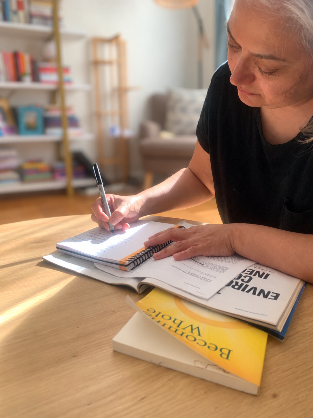 Nidia Trujillo writing observations in a notebook at a table surrounded by books and papers.