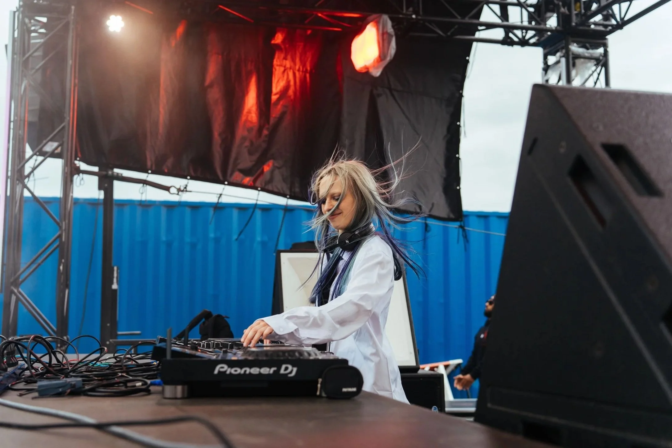 A female DJ with long hair, wearing a white shirt, spinning music at an outdoor event with DJ equipment on the table. There is a black sound system speaker in the foreground, and a stage with metal framework and orange lighting behind her. The backgr