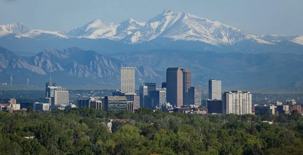 Denver city skyline with tall buildings in front of snow-capped mountains under a clear sky.