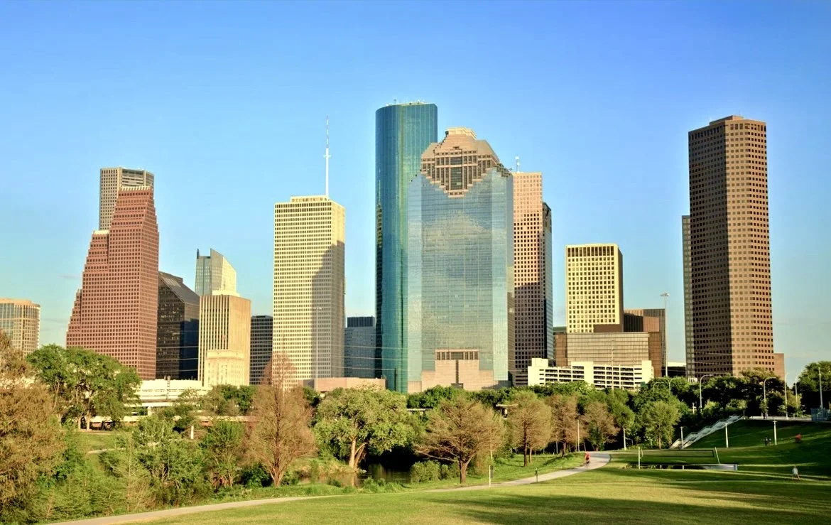 Skyline of downtown Houston, Texas, with tall office buildings and a park in the foreground under a clear blue sky.