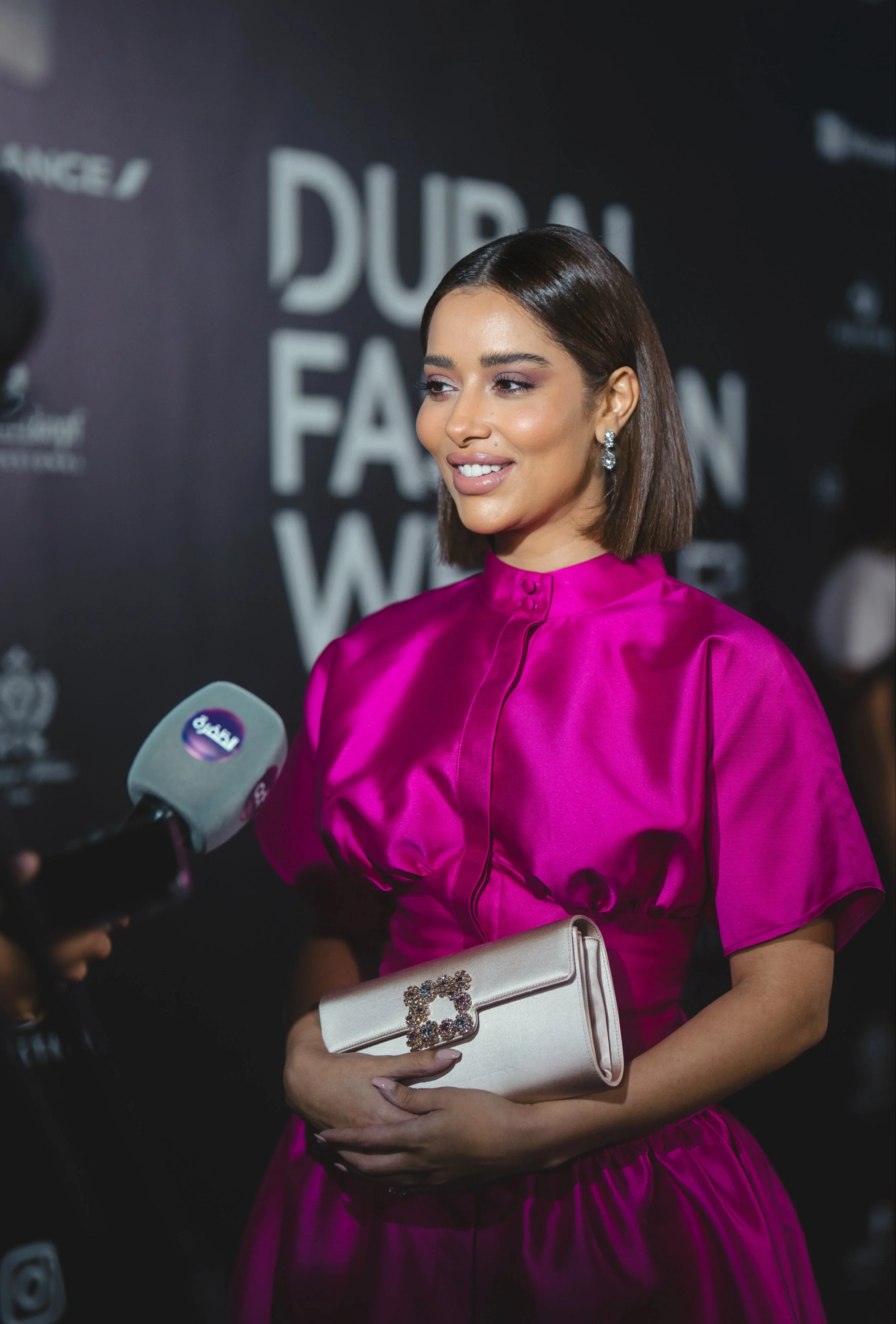 Une femme souriante portant une robe rose vif et tenant un sac à main crème avec une boucle décorative lors d'un événement fashion, avec un arrière-plan indiquant 'Dubai Fashion Week'.