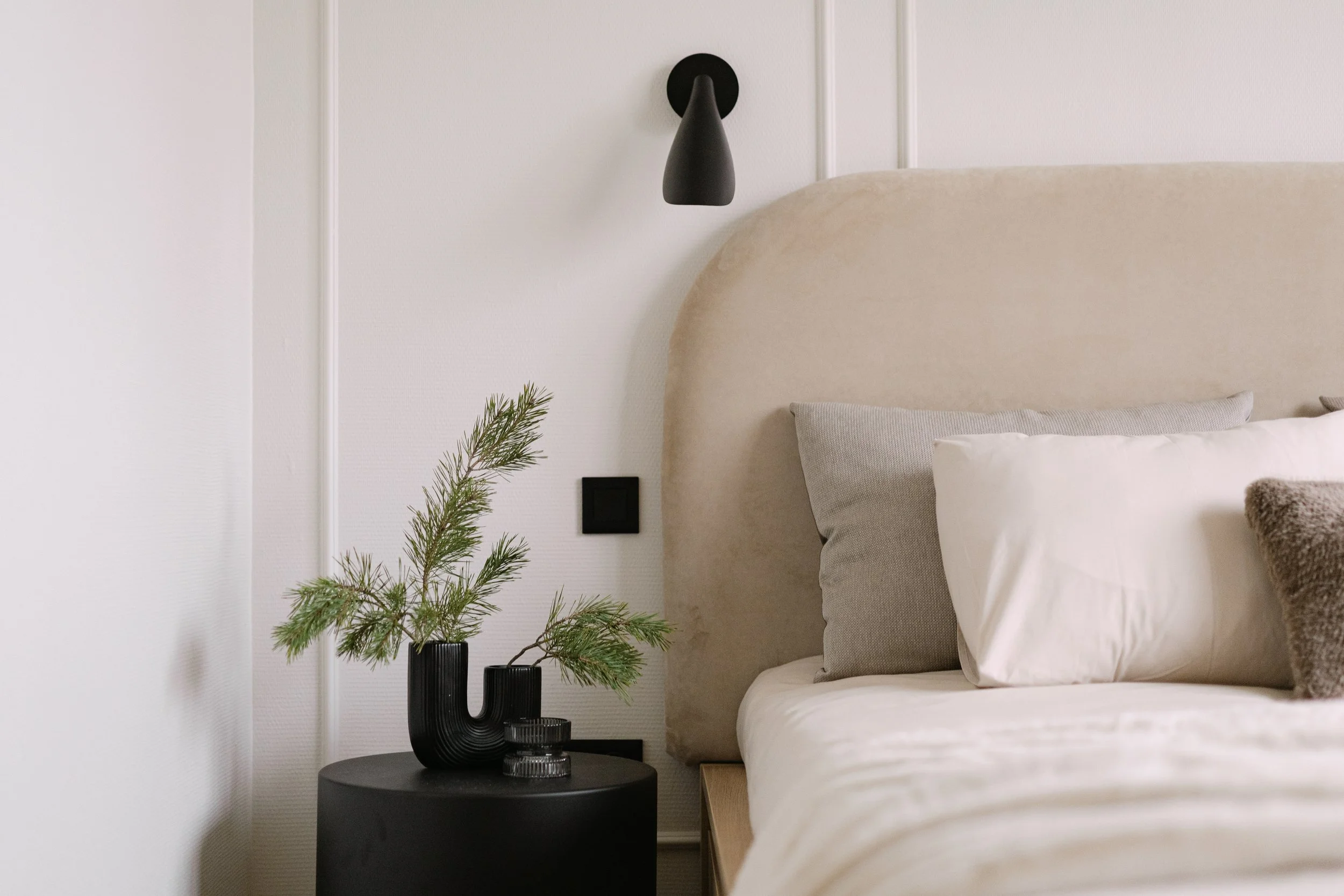 A minimalist bedroom with a beige upholstered bed, white pillow, and textured blanket. Next to the bed, there is a black round bedside table with a black vase holding green pine branches, and a black wall-mounted reading lamp above the bed.
