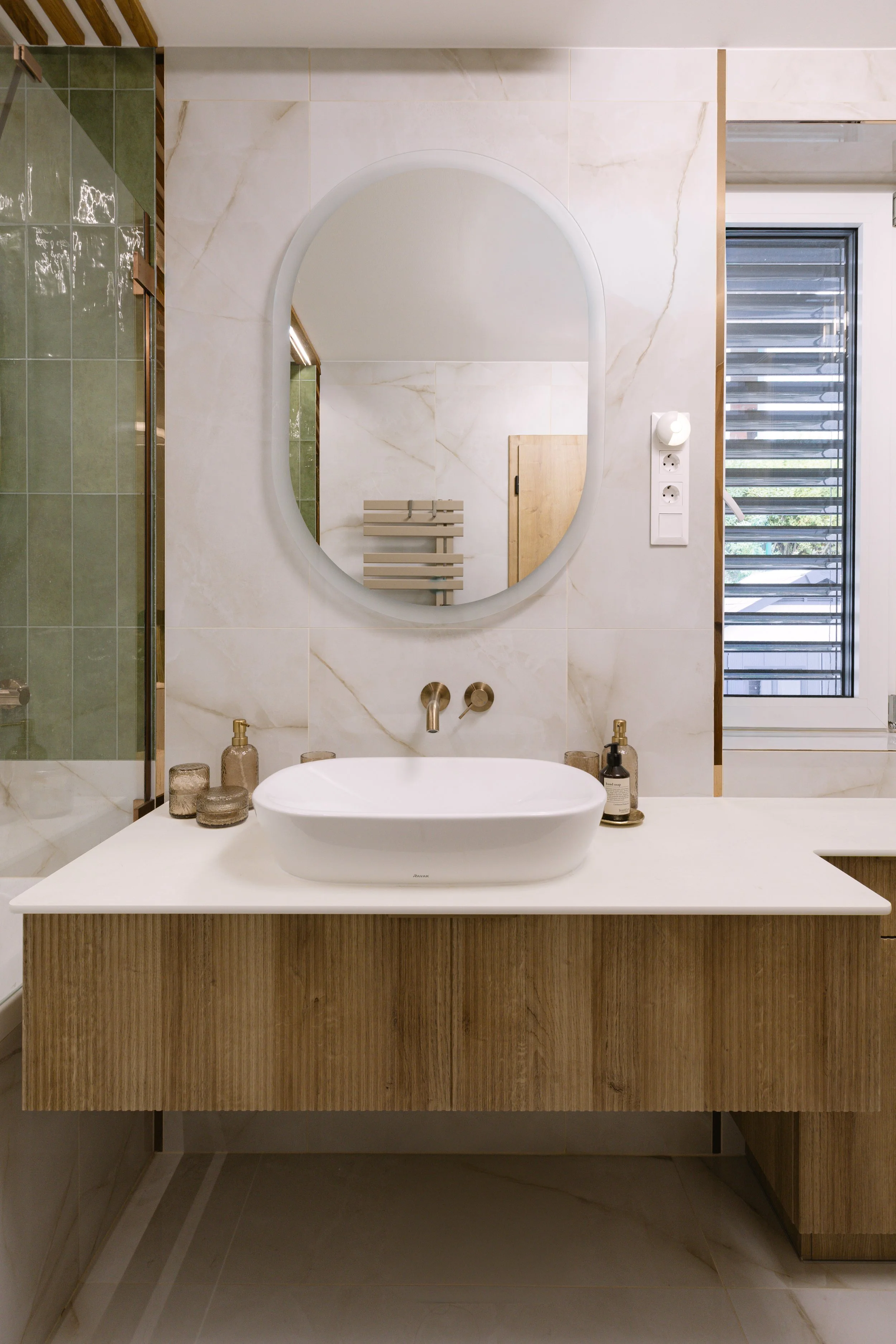 Modern bathroom with a rectangular ceramic sink, wooden vanity, round mirror, beige marble walls, and a window with horizontal blinds.