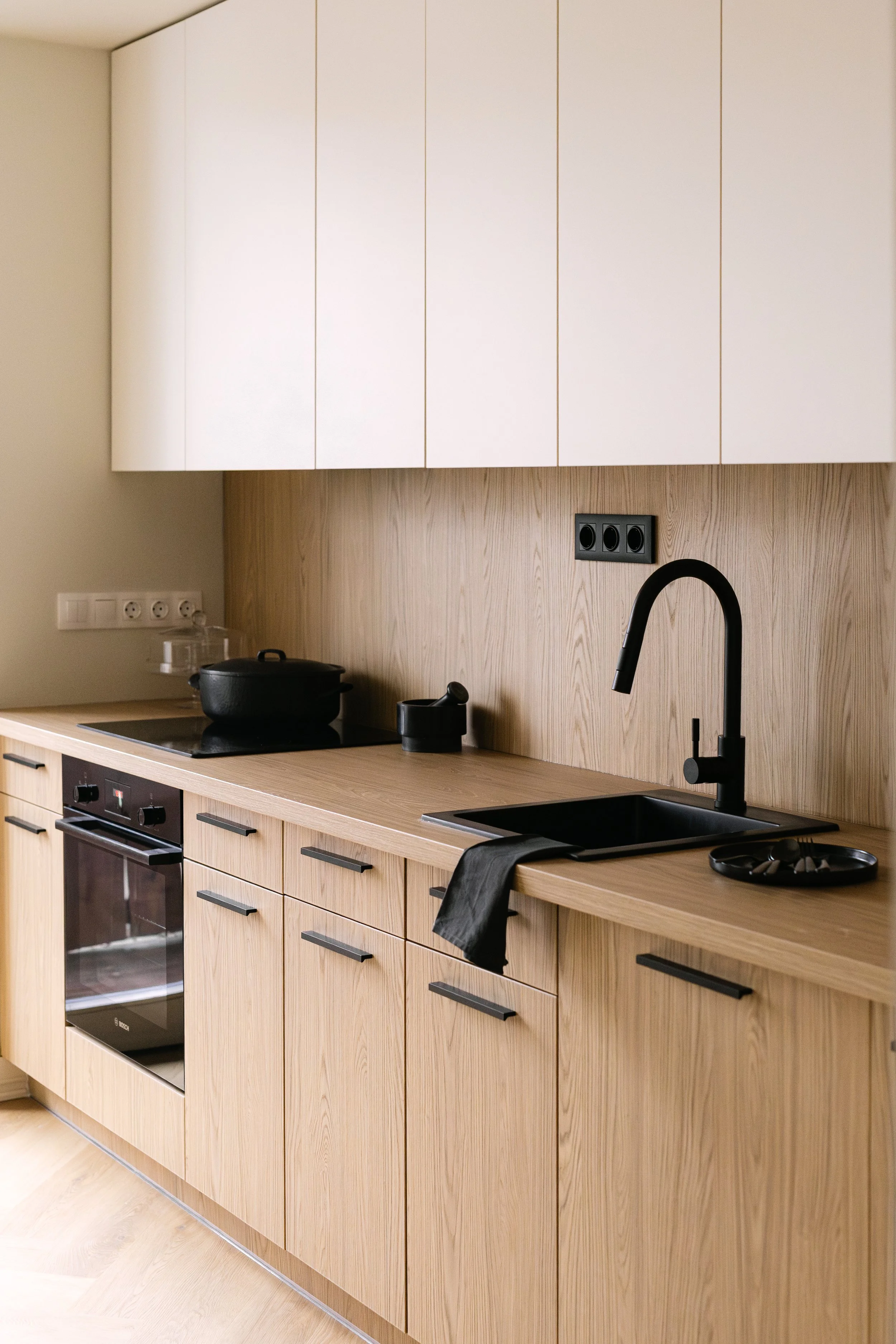 Modern kitchen with light wood cabinets, black stove, black sink with black faucet, and black utensils on the countertop. White wall with electrical outlets and a light wood backsplash.