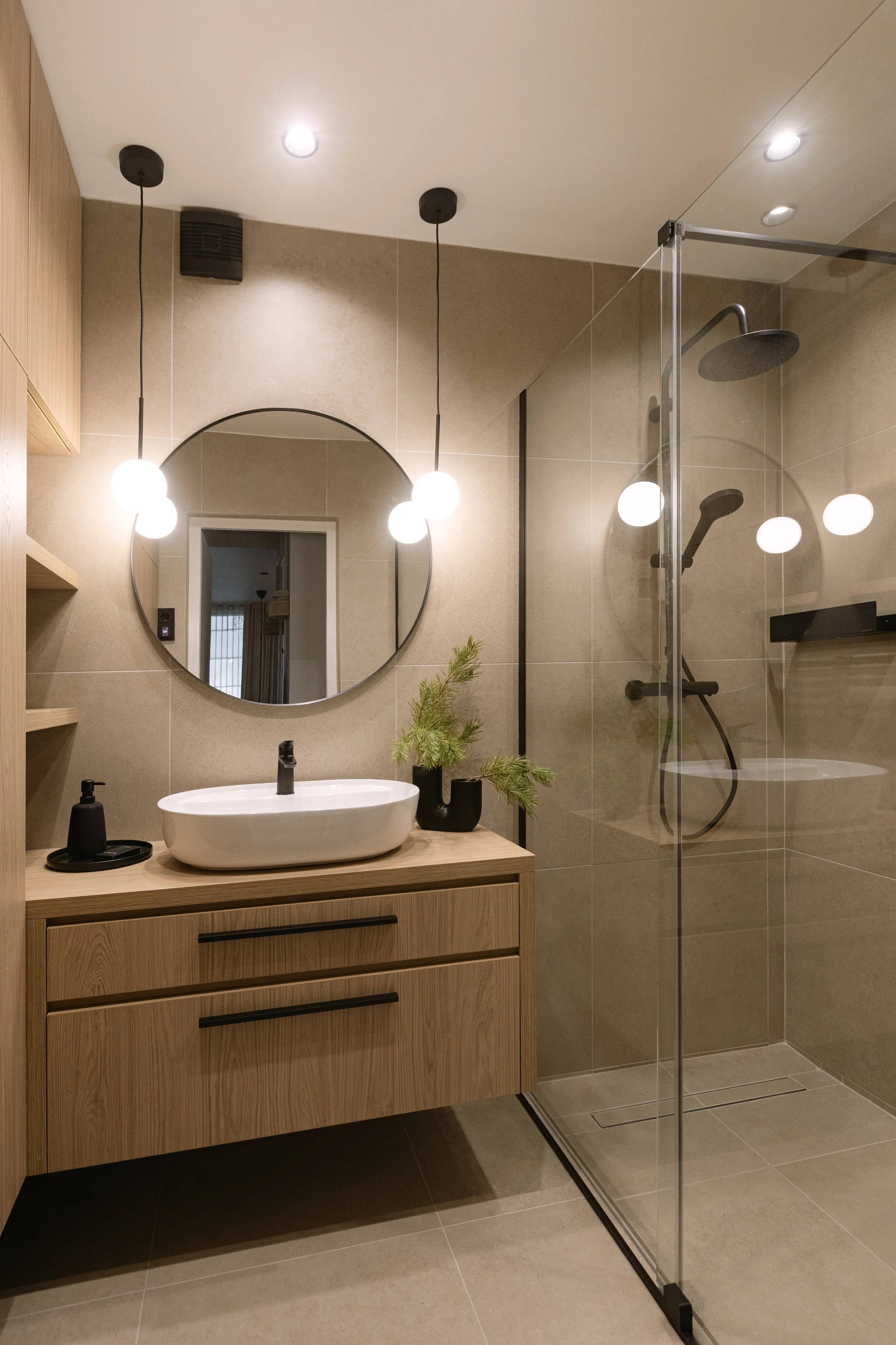 Modern bathroom features a wooden vanity with a white vessel sink, black faucet, and black soap dispensers. A round mirror hangs above the sink, and a small green plant decorates the countertop. To the right, a glass-enclosed shower has black fixture
