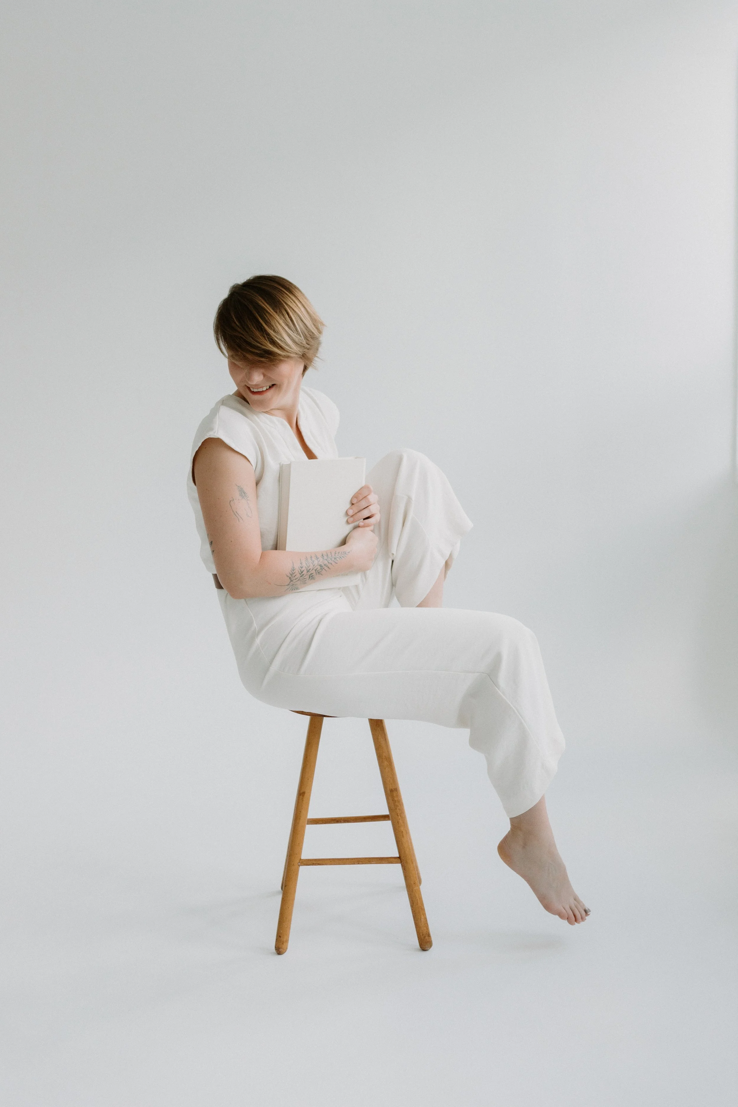 A woman with short brown hair smiling while sitting on a wooden stool, holding a white book and wearing a white outfit against a plain white background.