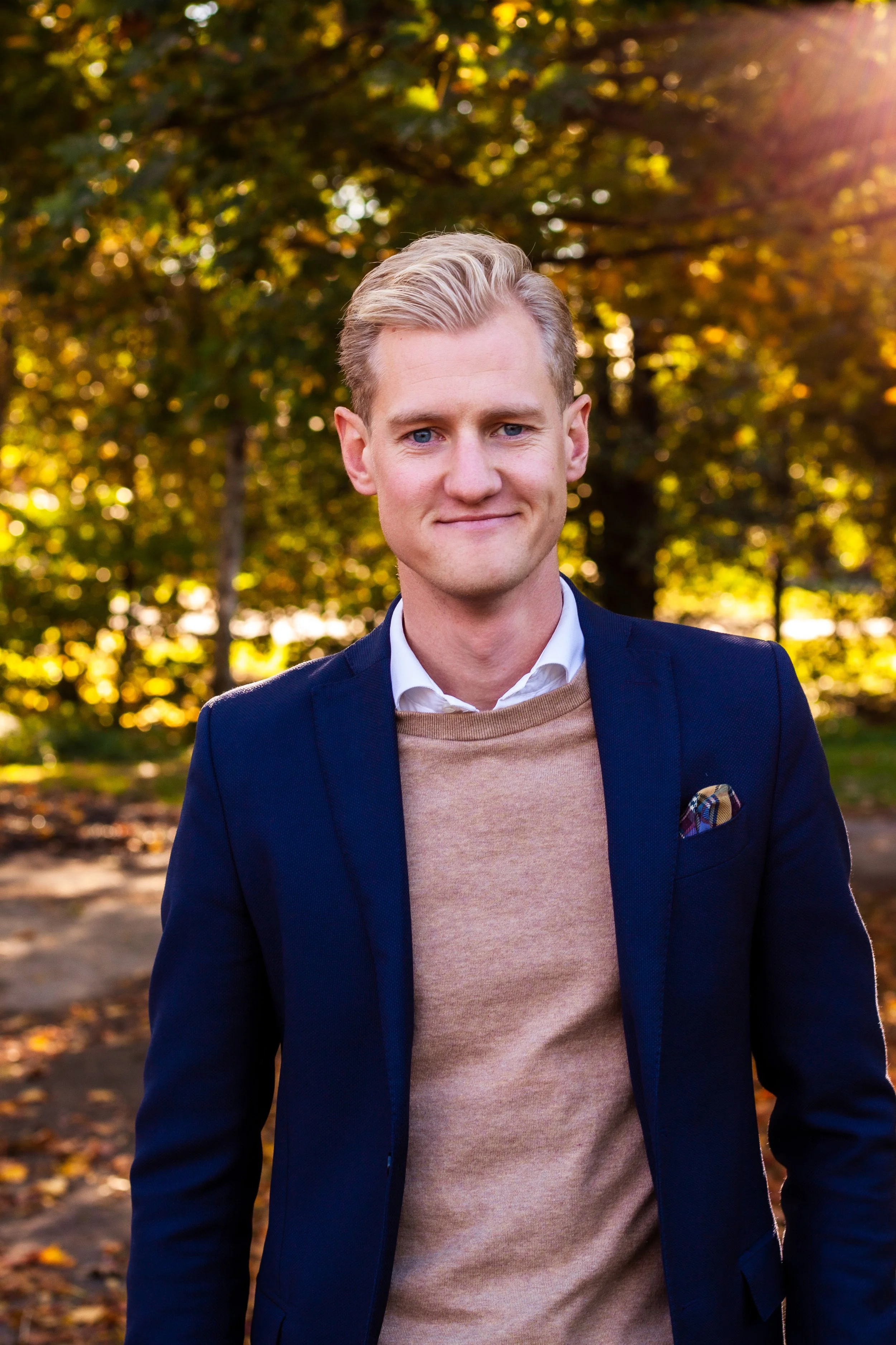 En ung mann i formell klær står utenfor med trær og høstfarger i bakgrunnen.