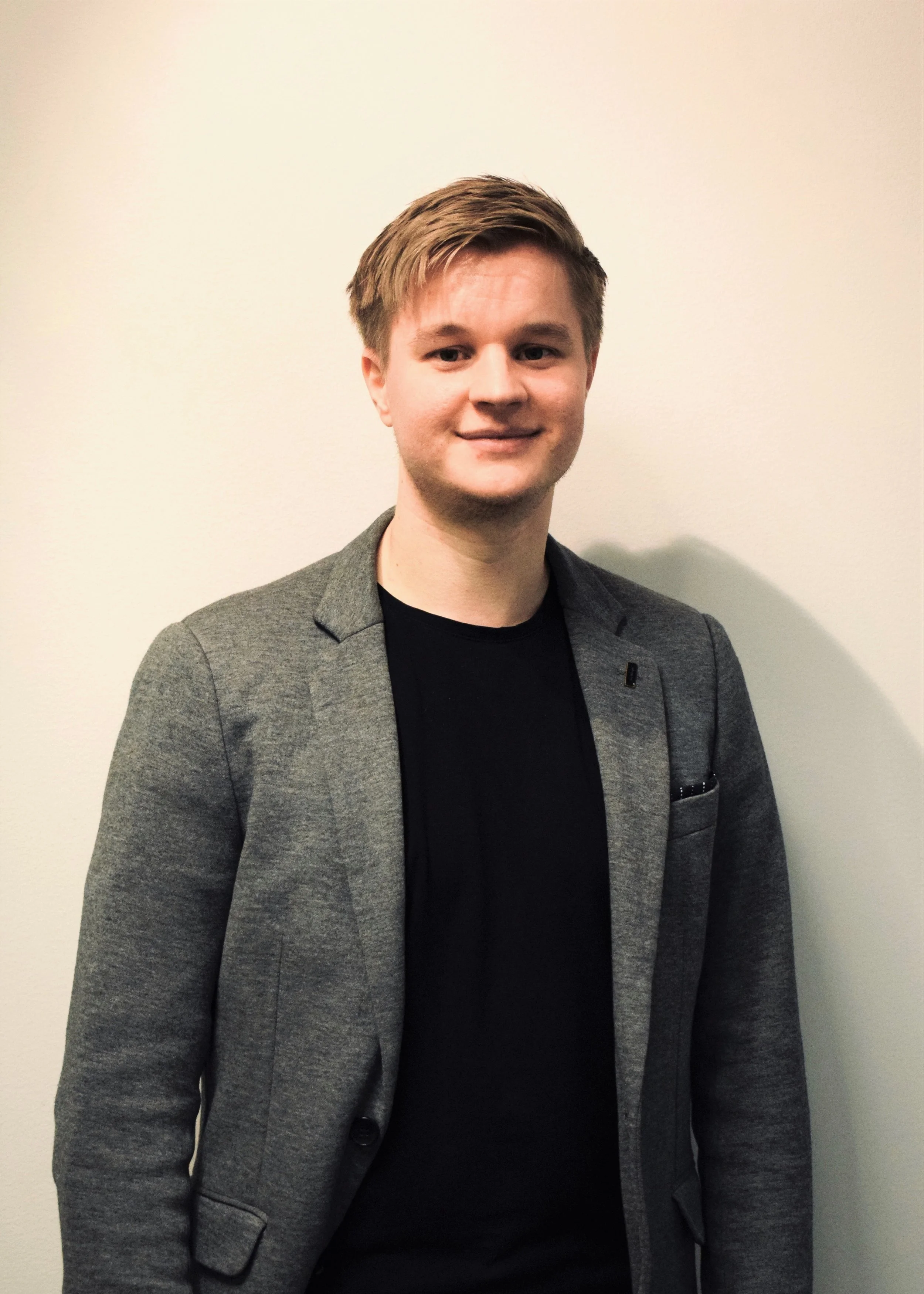 Ung mann med lett hår, kledd i en svart T-skjorte og en grå blazer, står mot en lys bakgrunn.
