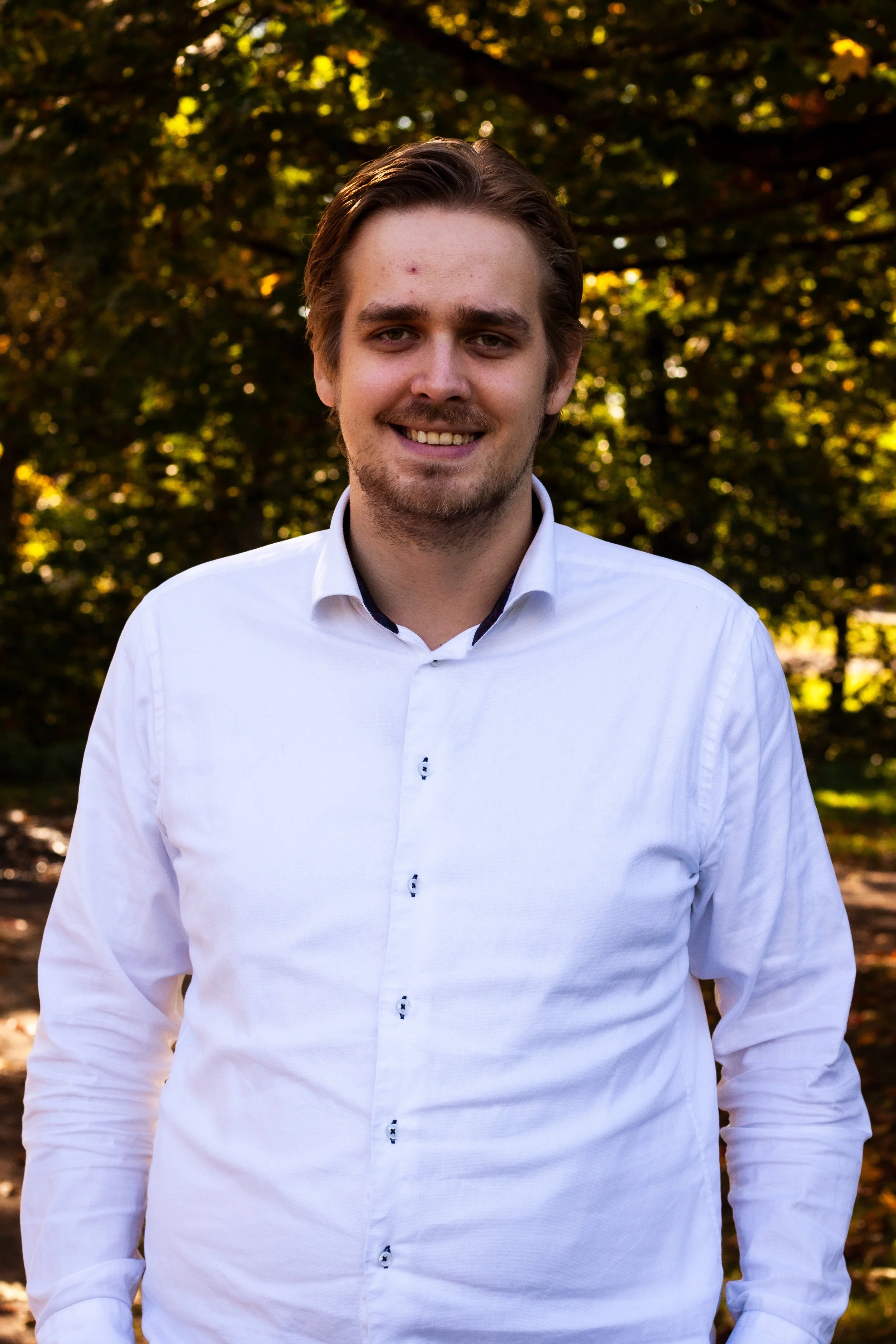 En ung mann med brunt, litt bustete hår, smilende, iført hvit skjorte, står utendørs foran trær med grønn og gul løvverk.