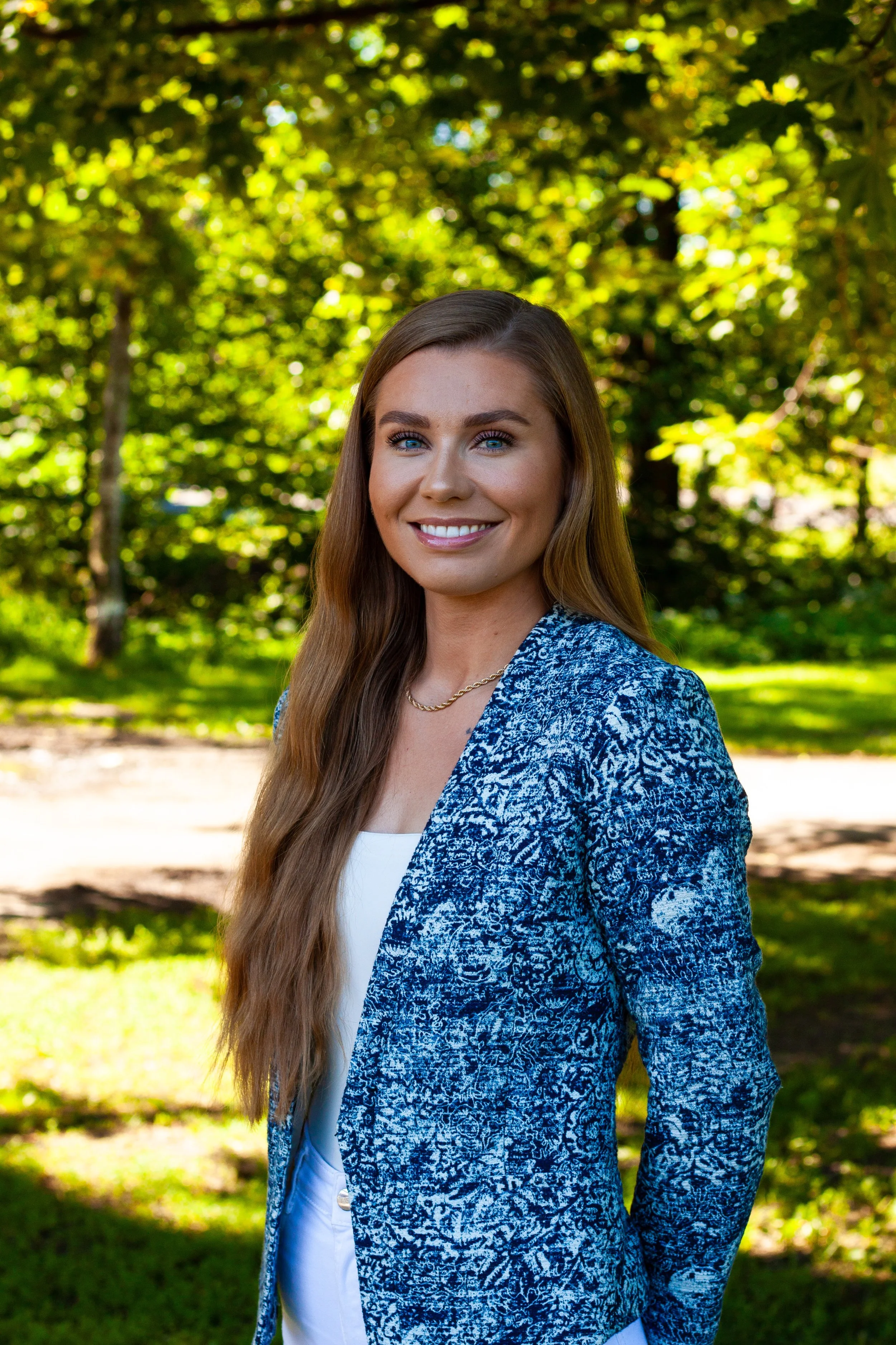 Portrett av en kvinne i en blå genserjakke med en hvit topp under, foran grønne planter.