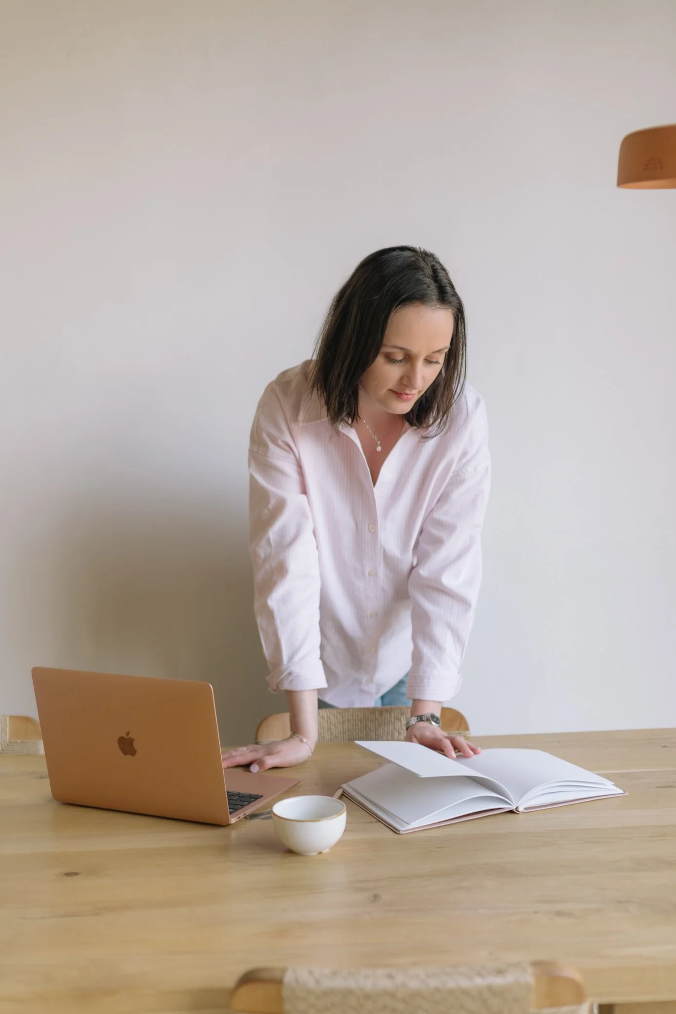 Femme avec des cheveux bruns courts portant une chemise rose clair, debout à côté d'une table en bois clair, regardant vers le bas avec un visage concentré. Sur la table, il y a un ordinateur portable rose doré, une tasse blanche, un livre ouvert et une feuille. Le fond est un mur blanc et une lampe beige suspendue.