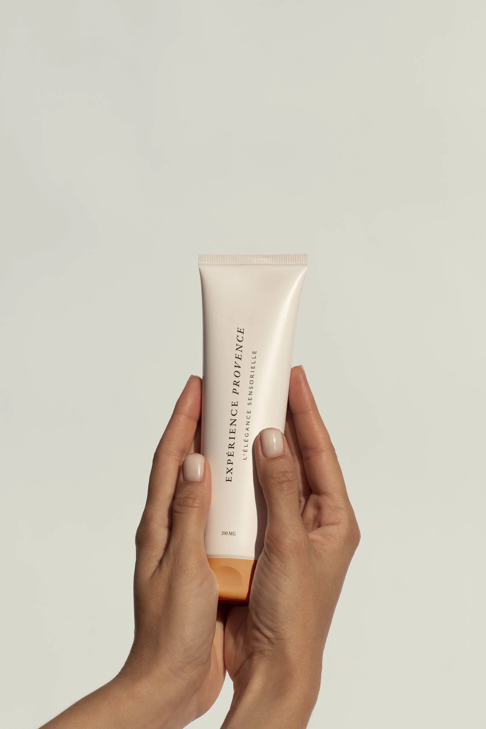 Une personne tient un tube blanc avec des mains. Le tube porte des inscriptions en français, notamment "EXPERIENCE", "L'ÉLÉGANCE PROVENÇALE" et "200 MG".