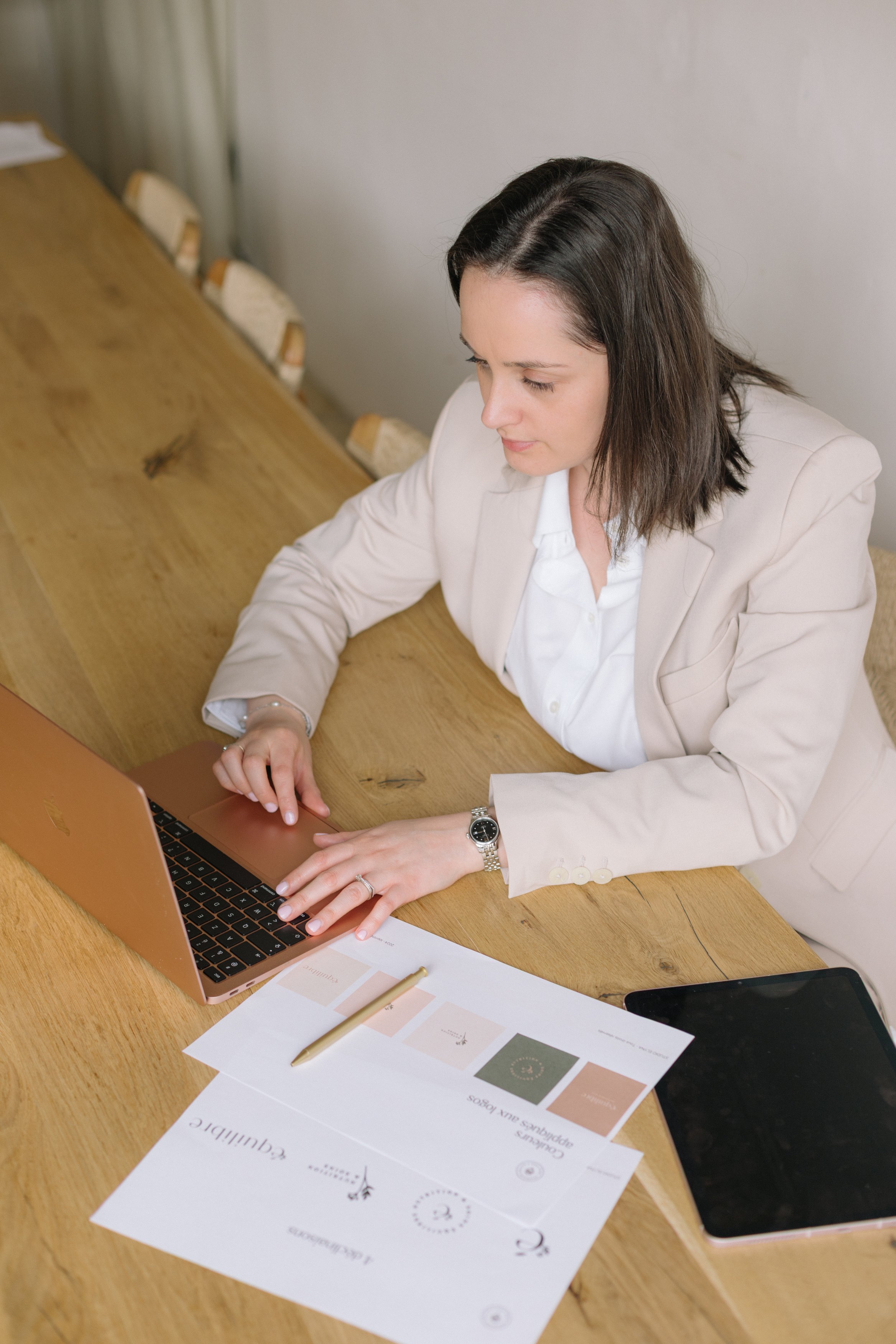 Une femme d'affaires assise à une table en bois, utilisant un ordinateur portable rose. Des papiers avec des graphiques et un smartphone sont aussi sur la table.
