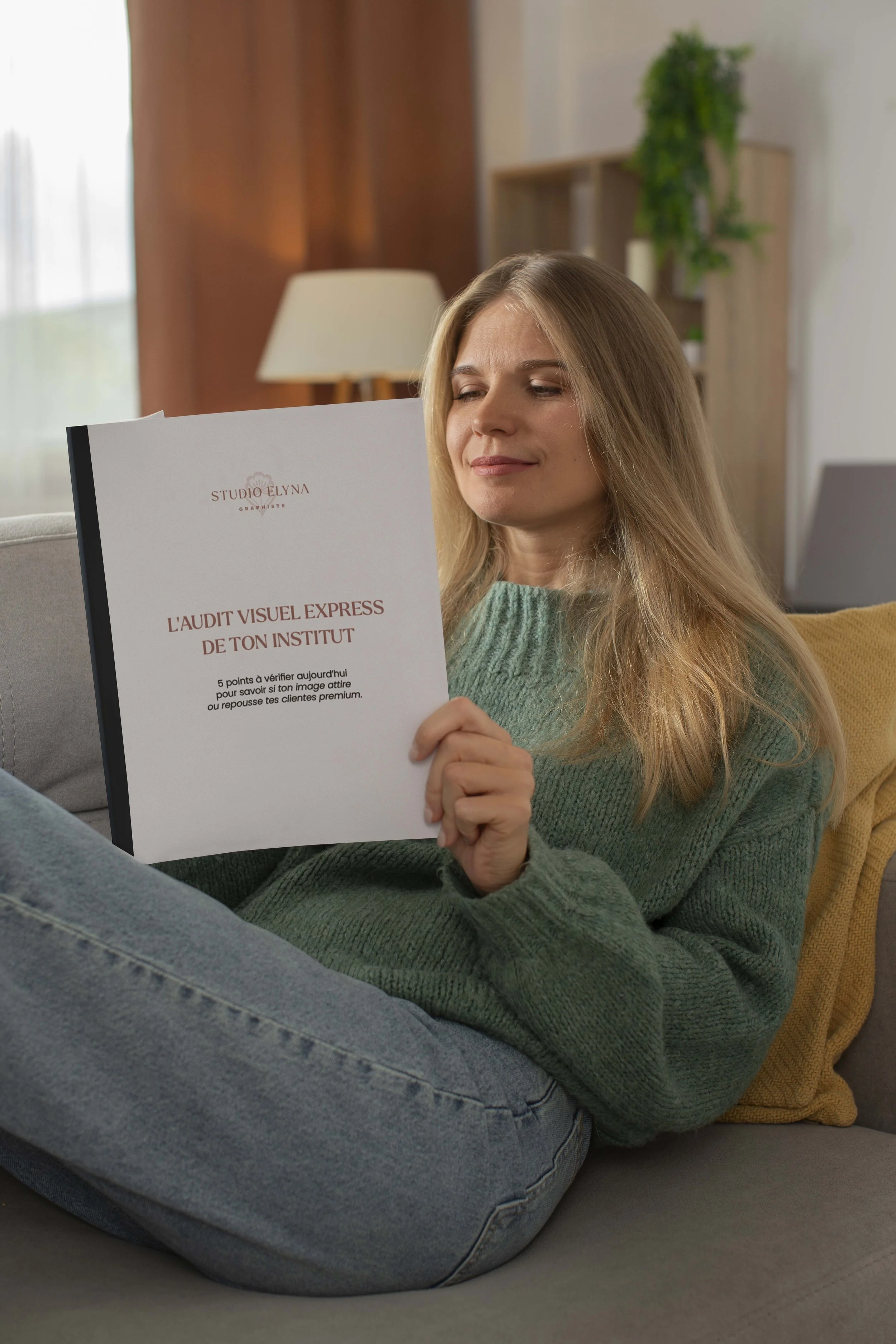 Une femme assise sur un canapé lit en train de regarder un document, dans un salon avec une lampe et des étagères en bois en arrière-plan.