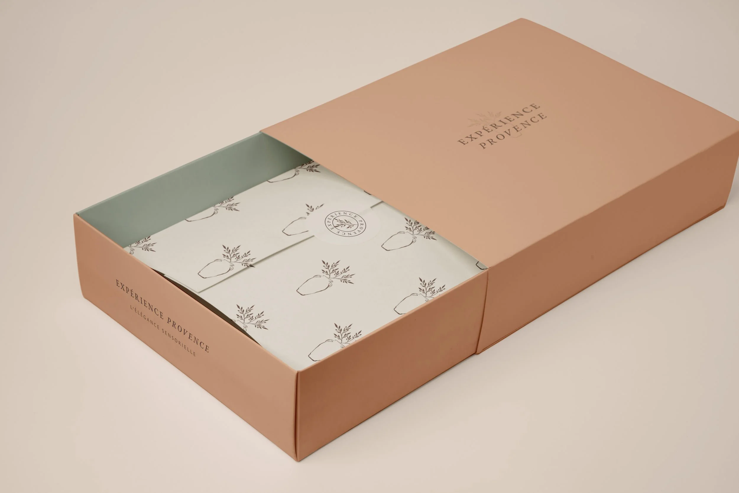 Une boîte rose avec un couvercle partiellement ouvert, contenant des papiers avec des illustrations de plantes et un logo "Expérience Provence".