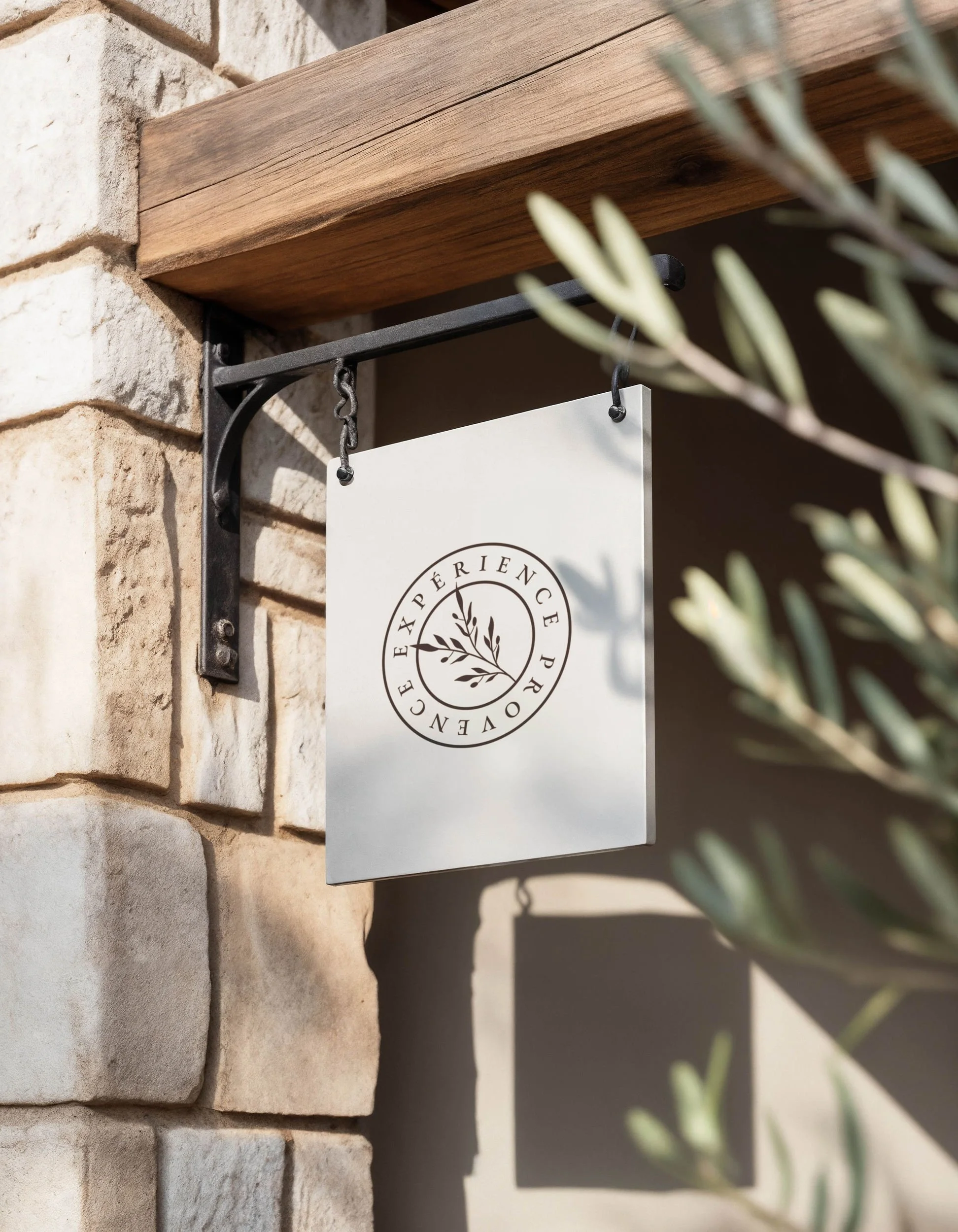 Un panneau blanc suspendu avec une boucle en haut, affichant un logo avec le texte 'EXPÉRIENCE PERENNE' et une branche d'olivier stylisée, fixé à un mur en pierre et toile de fond ombragée, avec des feuilles vertes en premier plan.