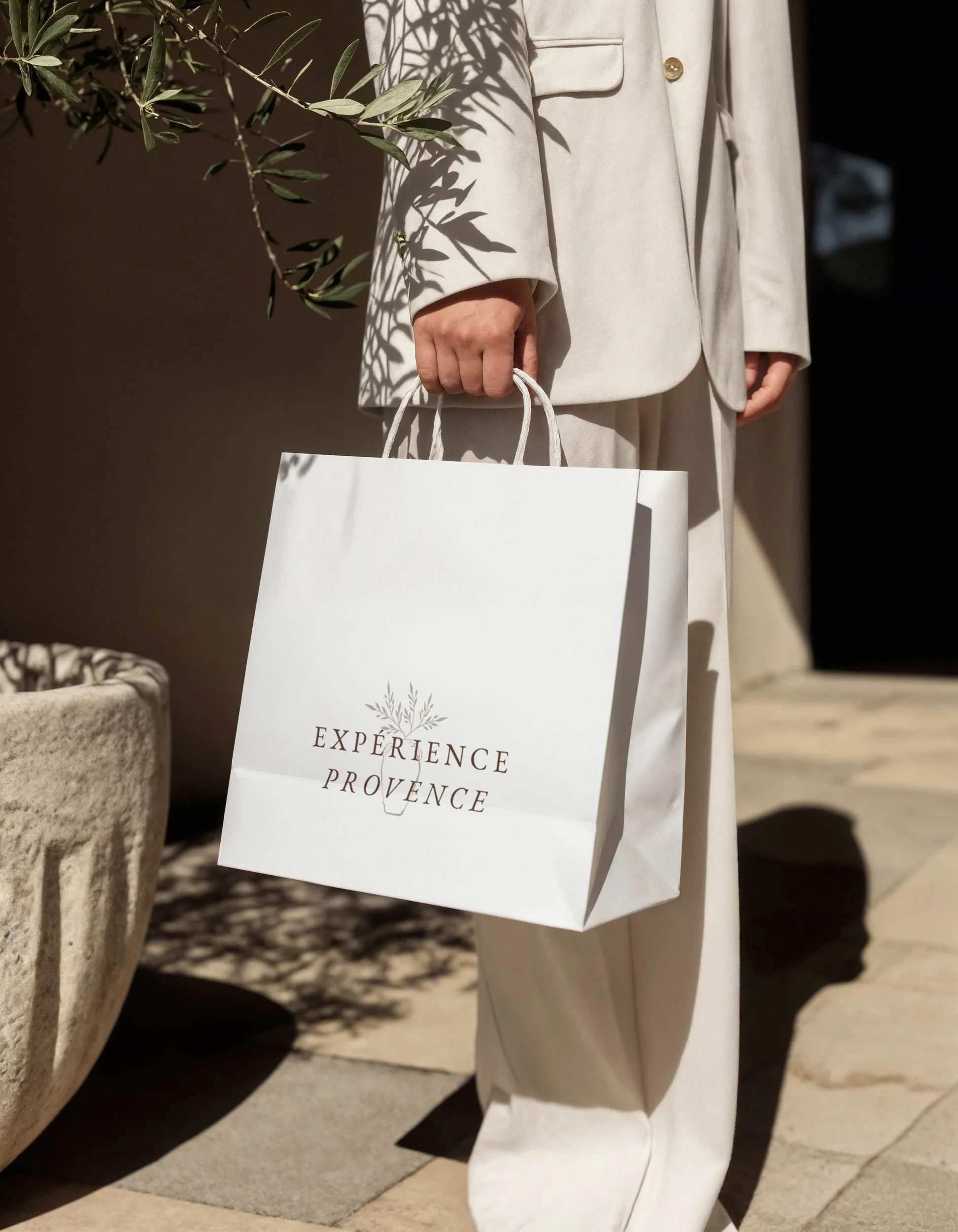 Personne portant un manteau beige et tenant un sac en papier blanc avec l'inscription "EXPÉRIENCE PROVENCE" dessus, en marchant sur un sol en pierre, avec un olivier en arrière-plan.
