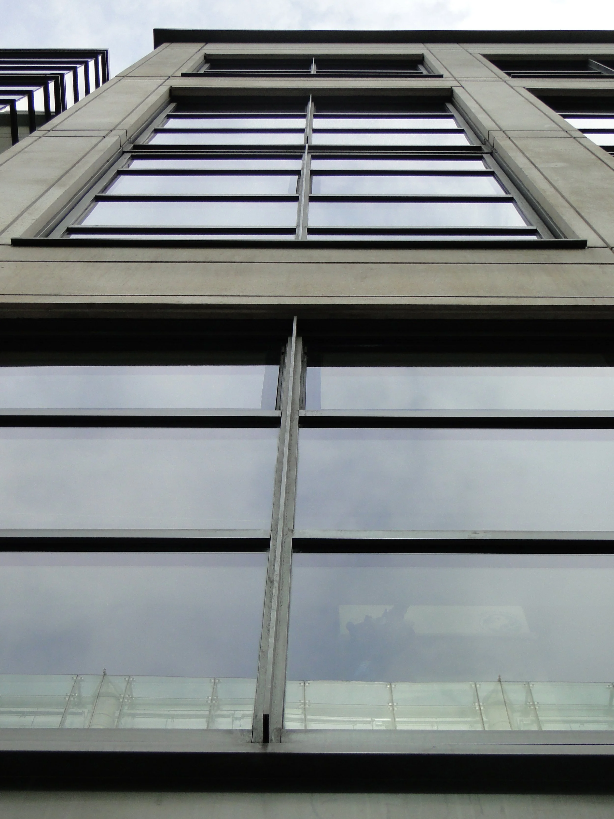 Low angle view of a modern multi-story building with large glass windows and concrete framing, overcast sky reflected in the windows.