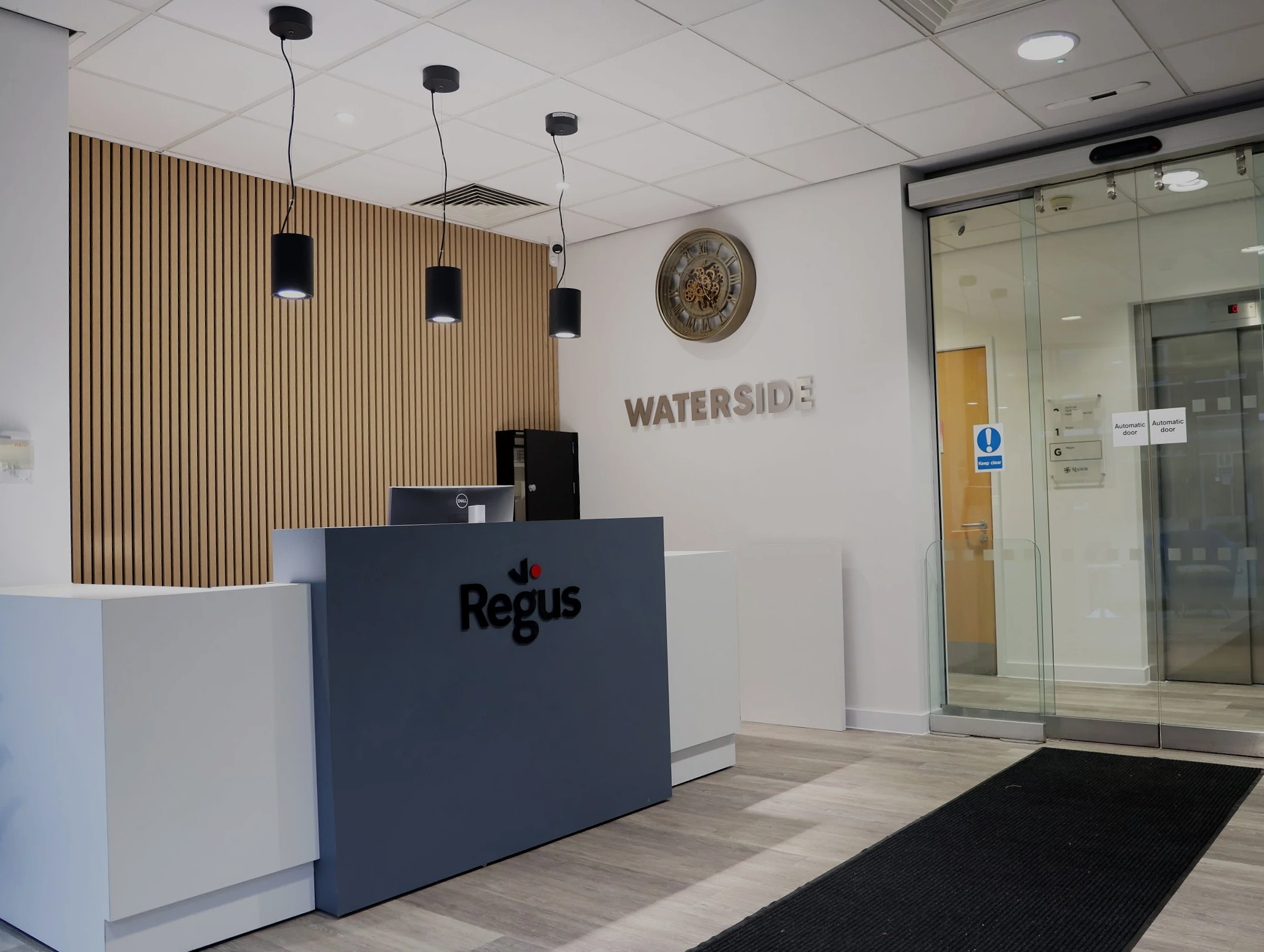 Reception desk with signage showing 'Regus' in a modern office lobby. Behind the desk, a wooden slat wall, a clock, and a sign that says 'WATERSIDE' are visible. There are black pendant lights hanging from the ceiling and a glass door to the right wi