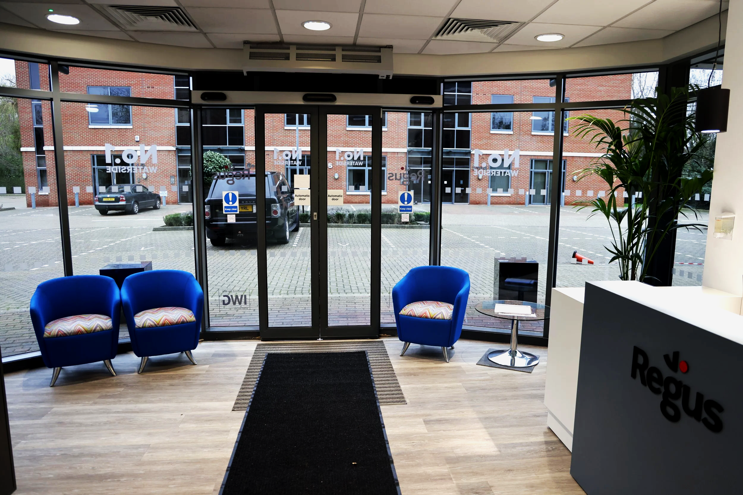 View of a modern office lobby with blue chairs and a small table, near a large glass entrance with a parking lot outside, and a sign reading 'Reclus' on a white reception desk on the right.