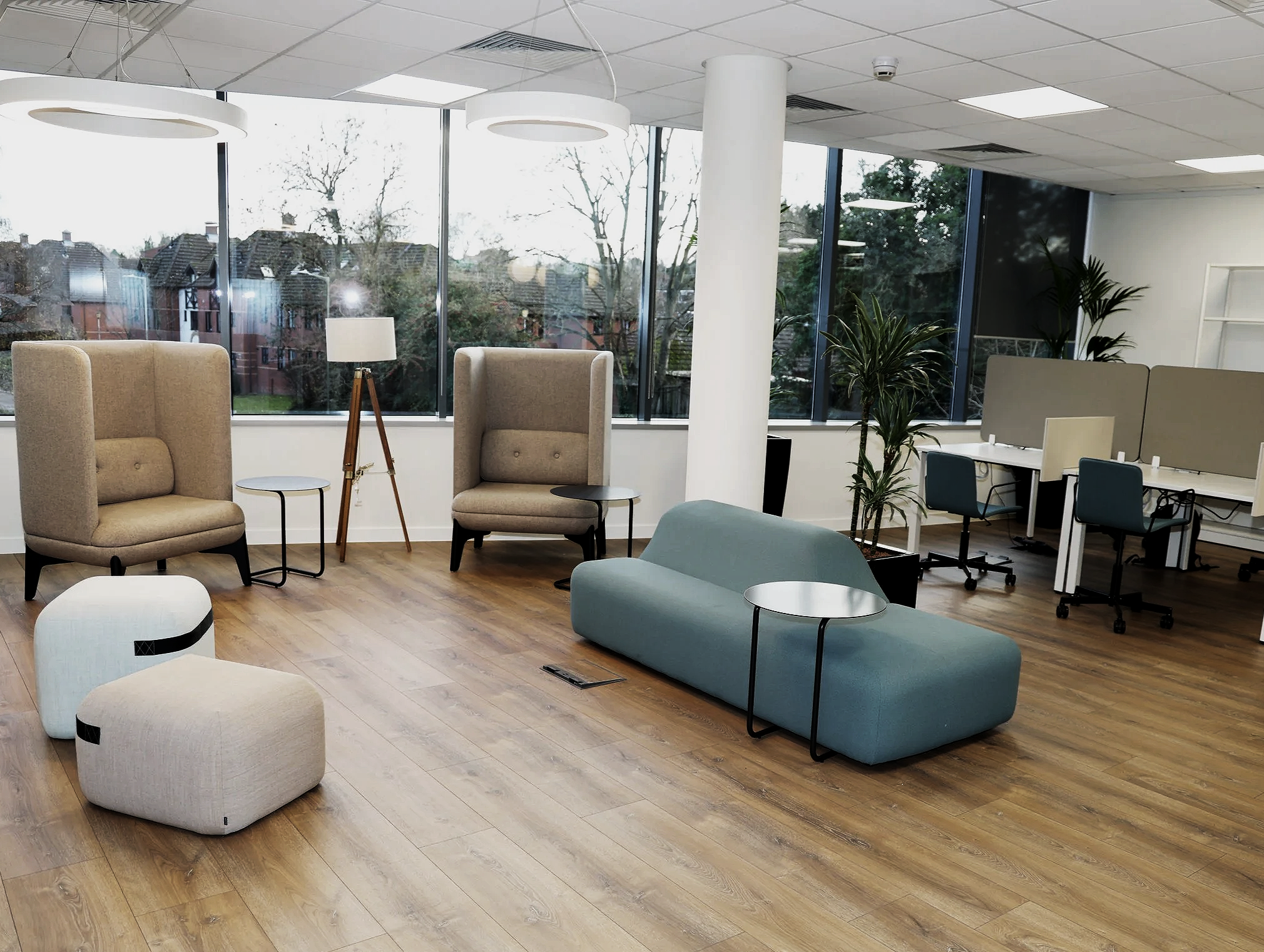 Modern office lounge with beige high-back chairs, small side tables, a blue sofa with a side table, and a work area with cubicles and chairs near large windows with trees outside.