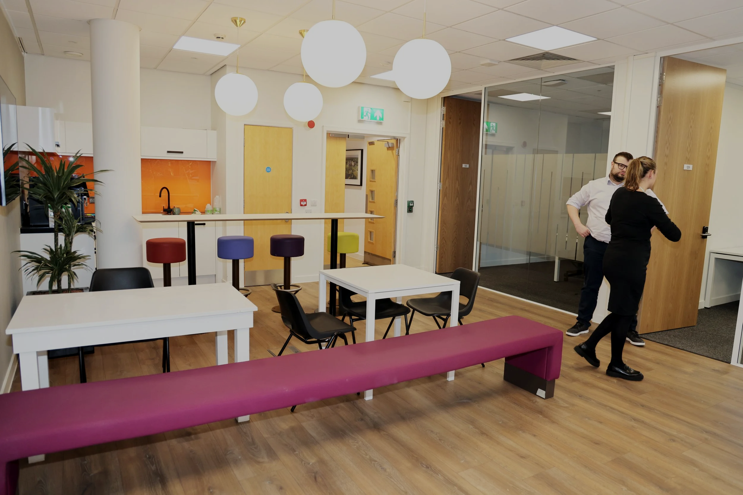Modern office lounge with white tables, black chairs, a pink bench, and a bar-height table with colorful stools. Two people are conversing near a wooden door, and the room features ceiling lights, a glass wall, and a kitchenette area.