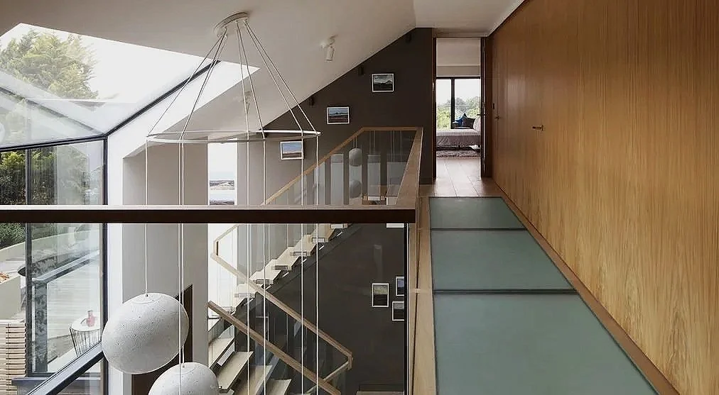 Interior view of a modern home with wood accents, glass railing, and a staircase, featuring large windows and minimalist decor.