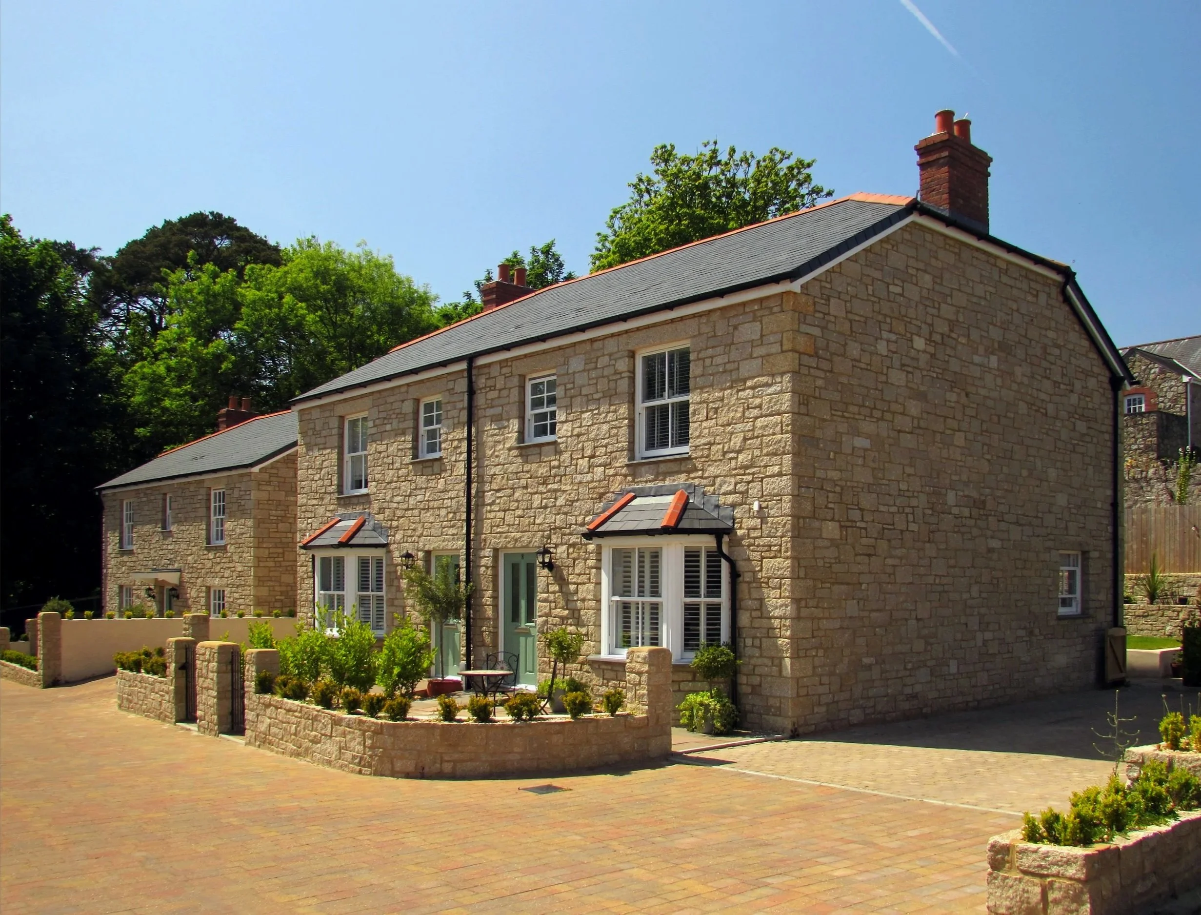 A stone-built two-story house with a dark gray roof, chimney, and front garden with plants and a small patio table.