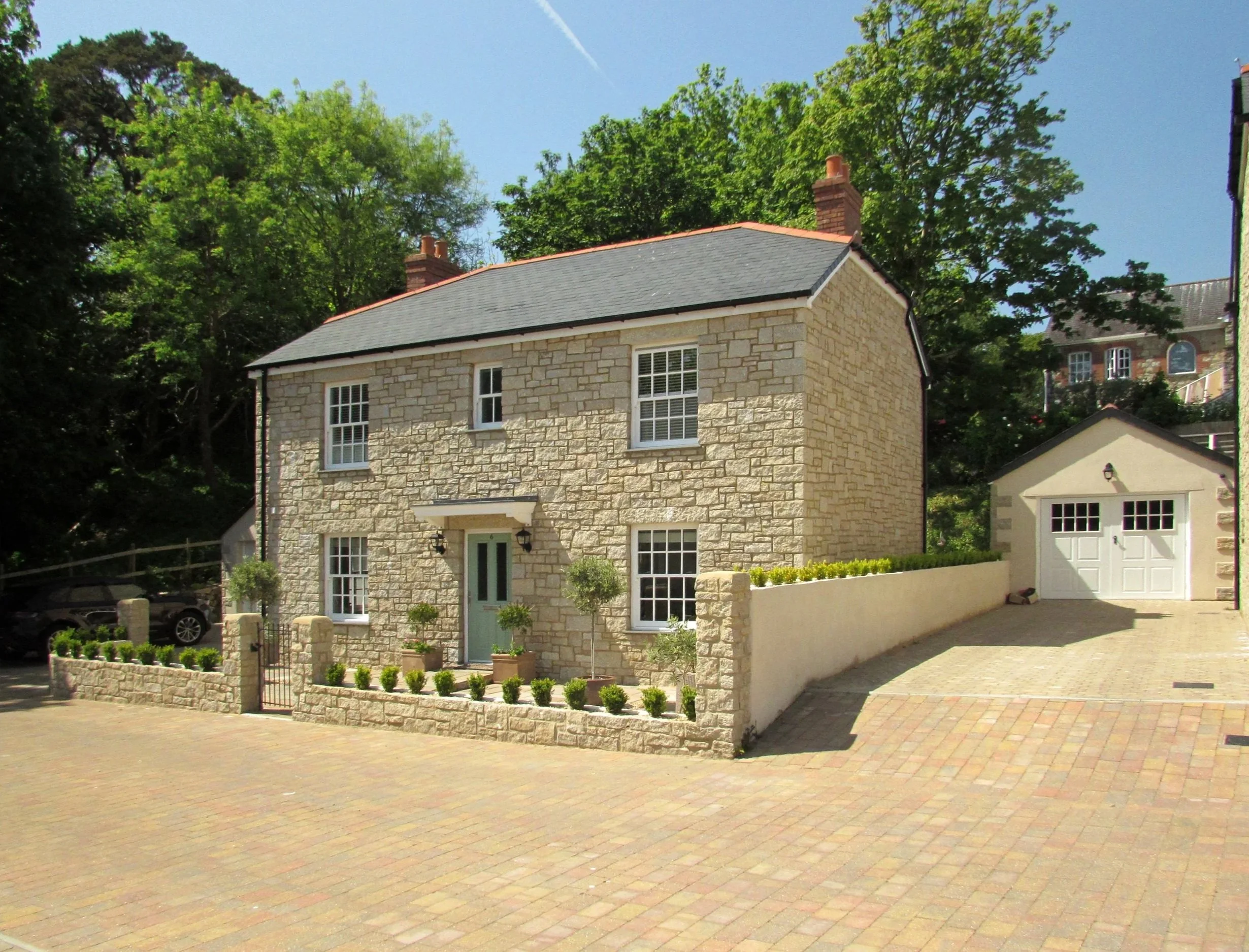 Stone house with a gray roof and white window frames, a small front garden with potted plants, and a driveway leading to a garage, under a blue sky with some clouds and green trees in the background.