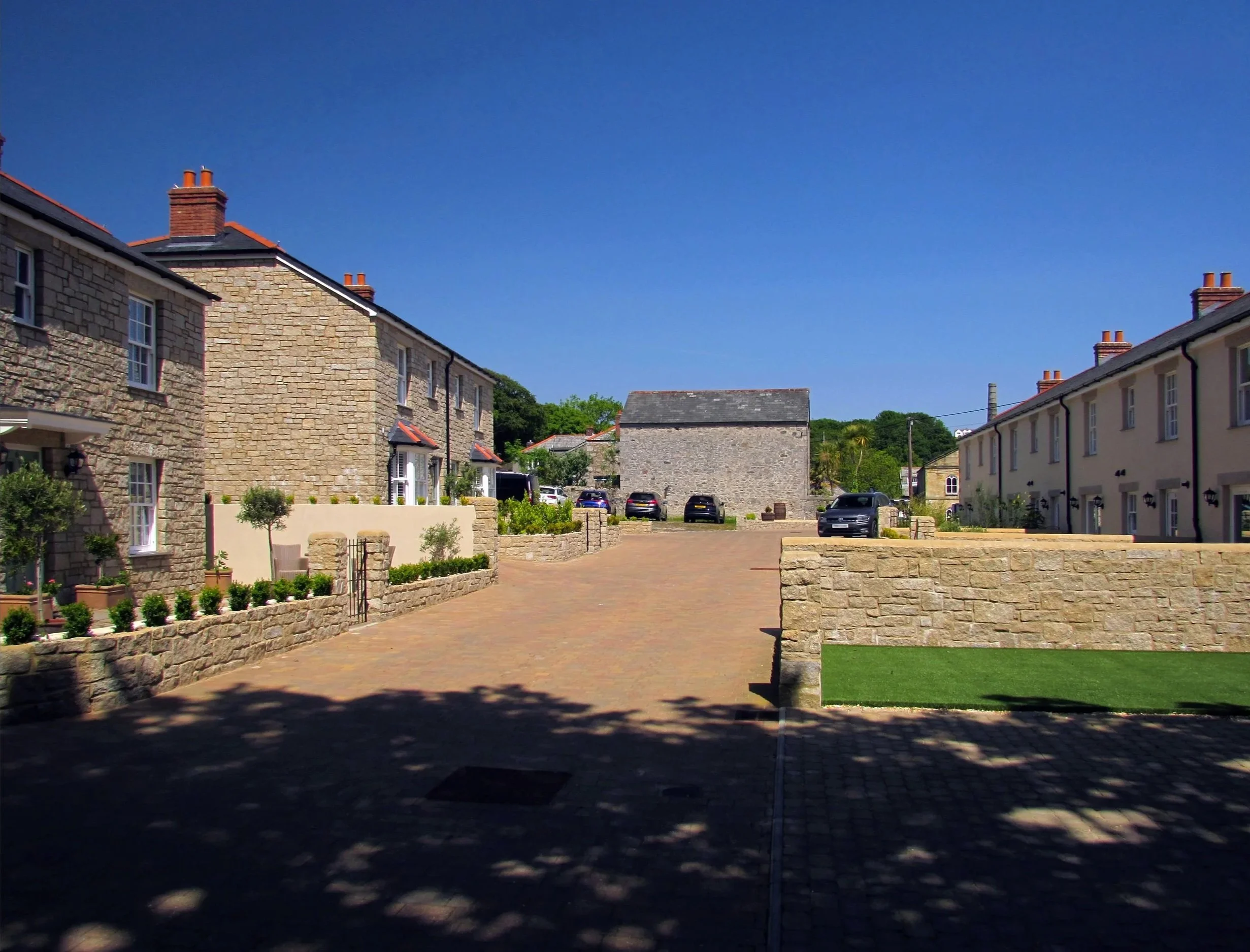 A residential street with stone houses on each side, a paved driveway in the middle, parked cars, and a clear blue sky.
