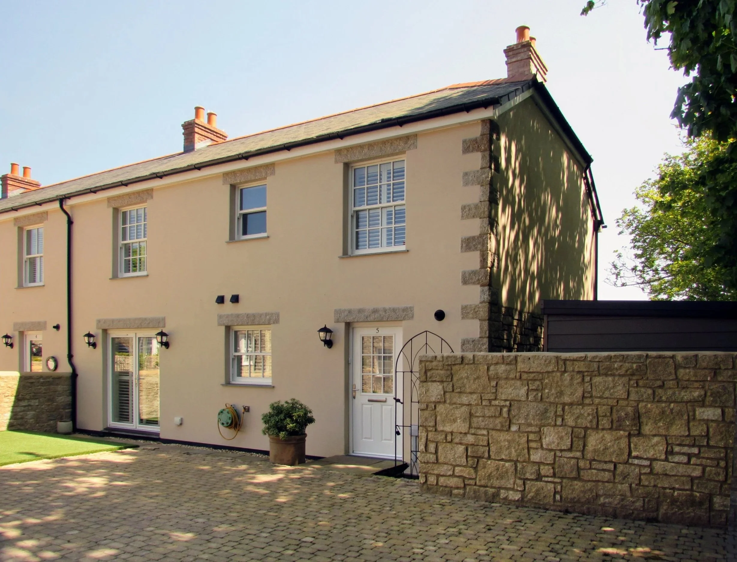 A cream-colored, two-story house with stone accents on the edges, surrounded by trees and a stone wall in the front yard. The house features white-framed windows, black outdoor wall lanterns, and a white door with a decorative metal archway. The yard