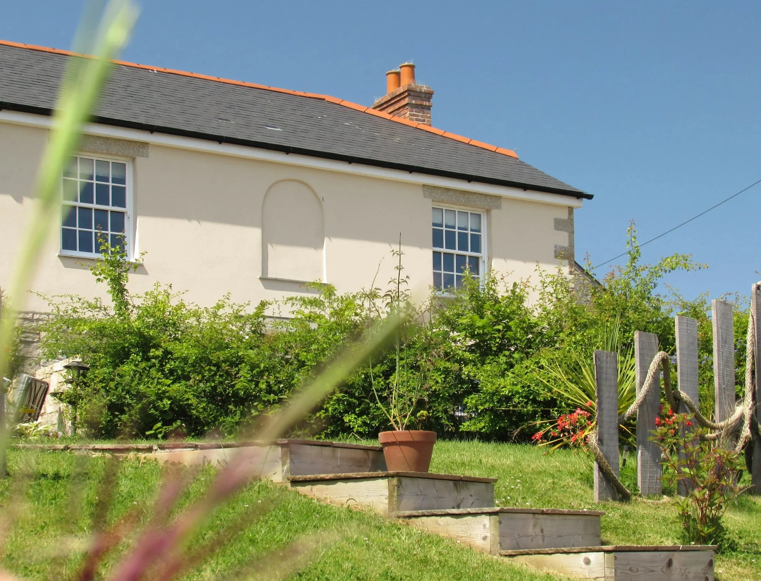 A white house with a gray roof and a brick chimney surrounded by a green garden with potted plants, shrubs, and a wooden fence, under a clear blue sky.