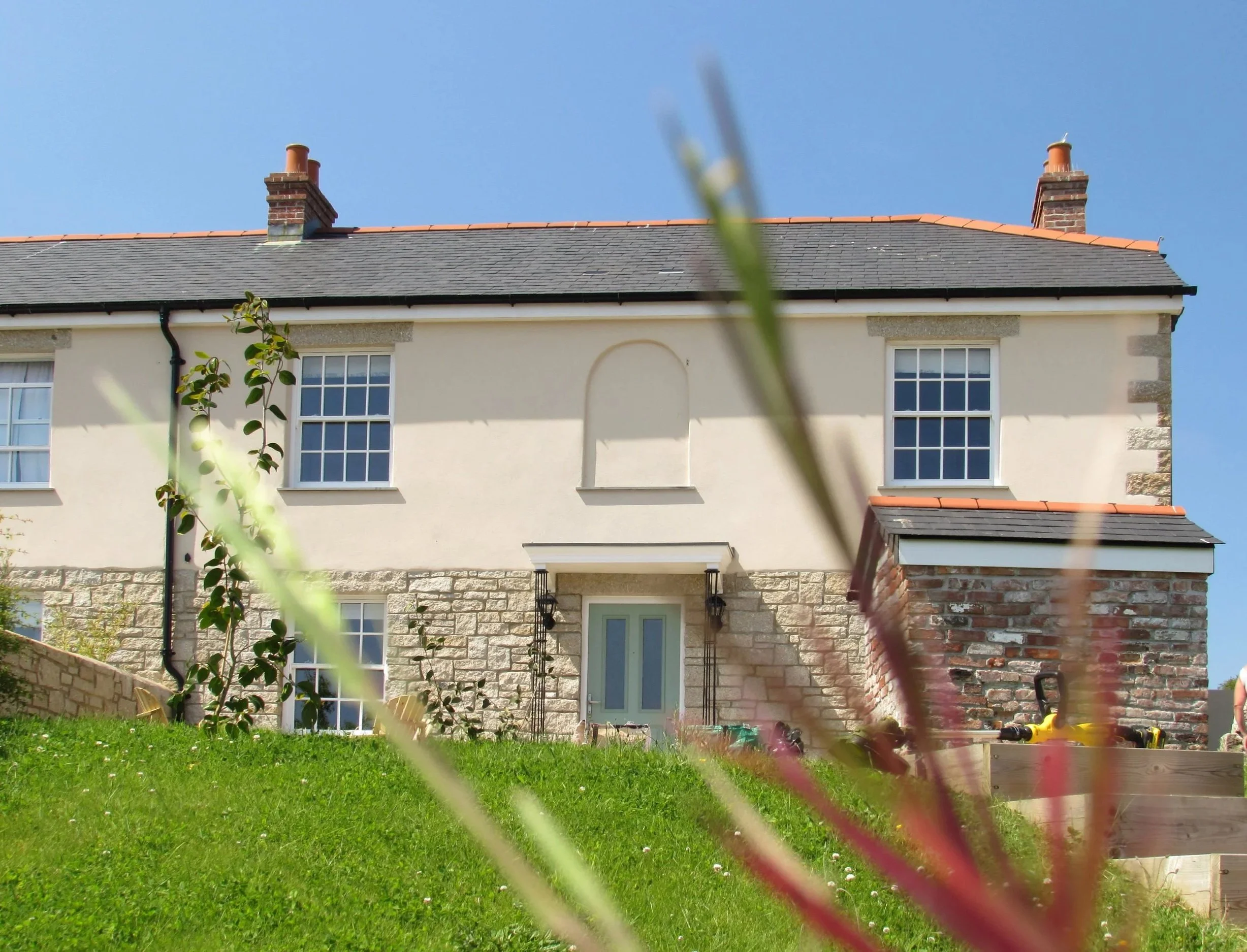 Front view of a two-story house with a stone and stucco exterior, three windows and a garage with roofing tiles, set on a grassy slope with plants and flowers in the foreground.