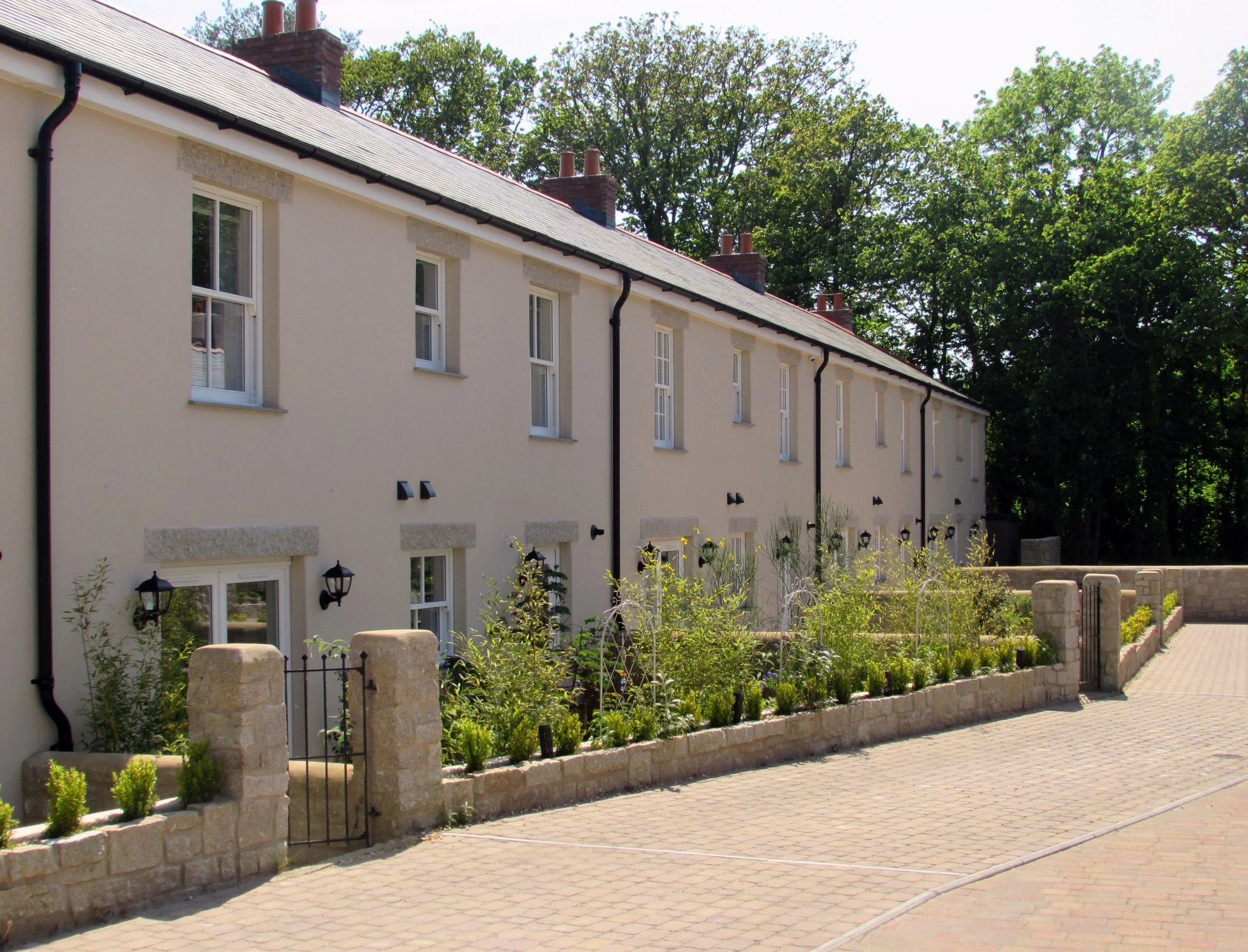 A row of white residential townhouses with black shutters and small front garden, enclosed by a stone wall and wrought iron gate, with a paved driveway in front and trees in the background.