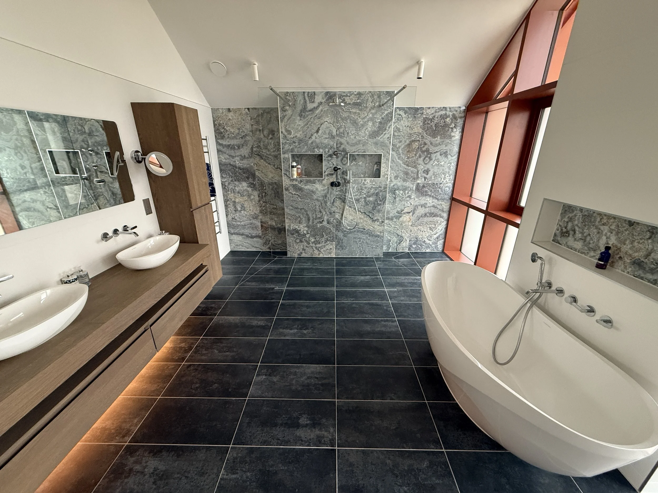Modern bathroom with black tiled floor, double sinks on wooden vanity, large mirror, walk-in shower with gray stone wall, and free-standing bathtub near red window frames.