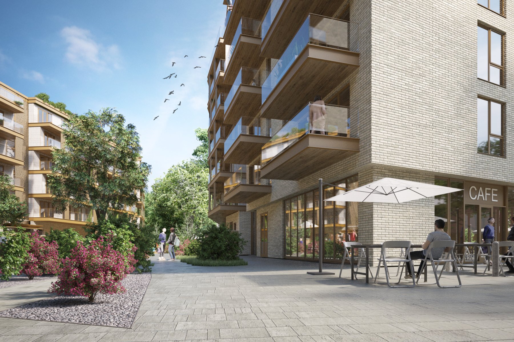 A modern residential complex with a cafe on the ground floor. Outdoor seating with tables, chairs, and a large white umbrella. People are sitting and walking around in a landscaped area with trees and pink flowering shrubs, under a blue sky with some