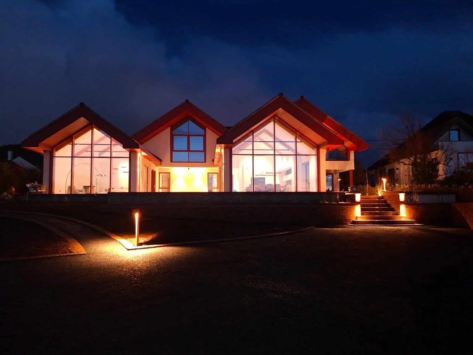 Modern house at night with large glass windows illuminated from inside, stairs with lights leading to the entrance, dark cloudy sky in the background.