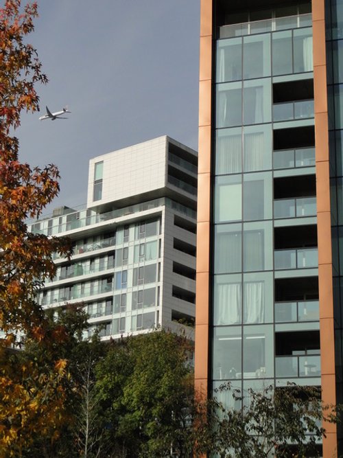 View of modern high-rise apartment buildings with glass balconies, trees in the foreground, and an airplane flying in the sky.